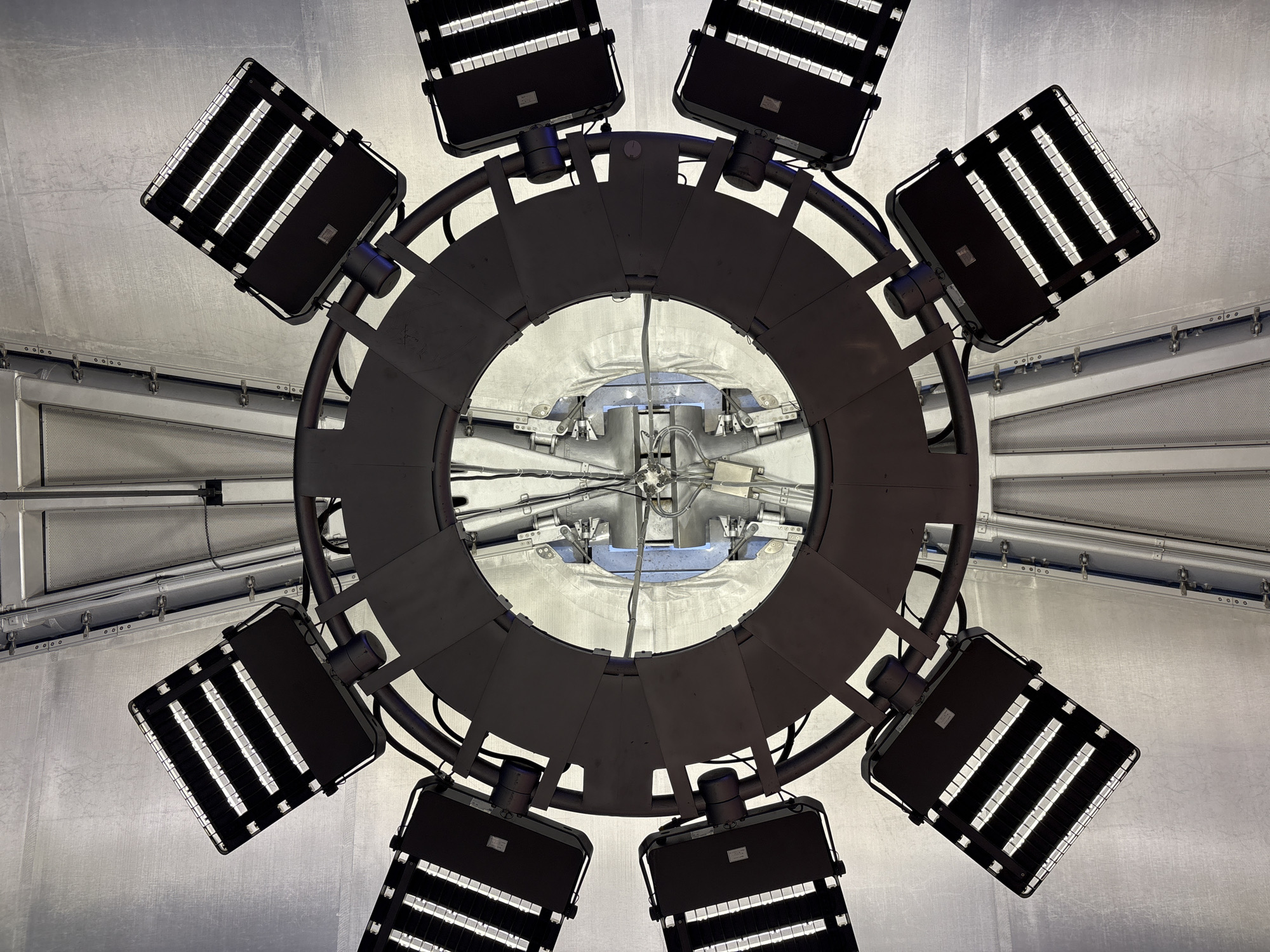 An overhead perspective captures a circular arrangement of eight black floodlights mounted on a dark metallic ring. The floodlights are angled outwards and upwards, their bright LED strips illuminating the surrounding metallic ceiling. The ceiling appears to be constructed of corrugated metal panels, with some sections featuring a perforated texture. In the center of the ring, a complex structure of wires, metal supports, and what appears to be part of a light fixture or mechanism is visible. The overall scene suggests an industrial or large-scale lighting installation, possibly in a stadium, arena, or warehouse. The location is described as Unknown, Unknown.