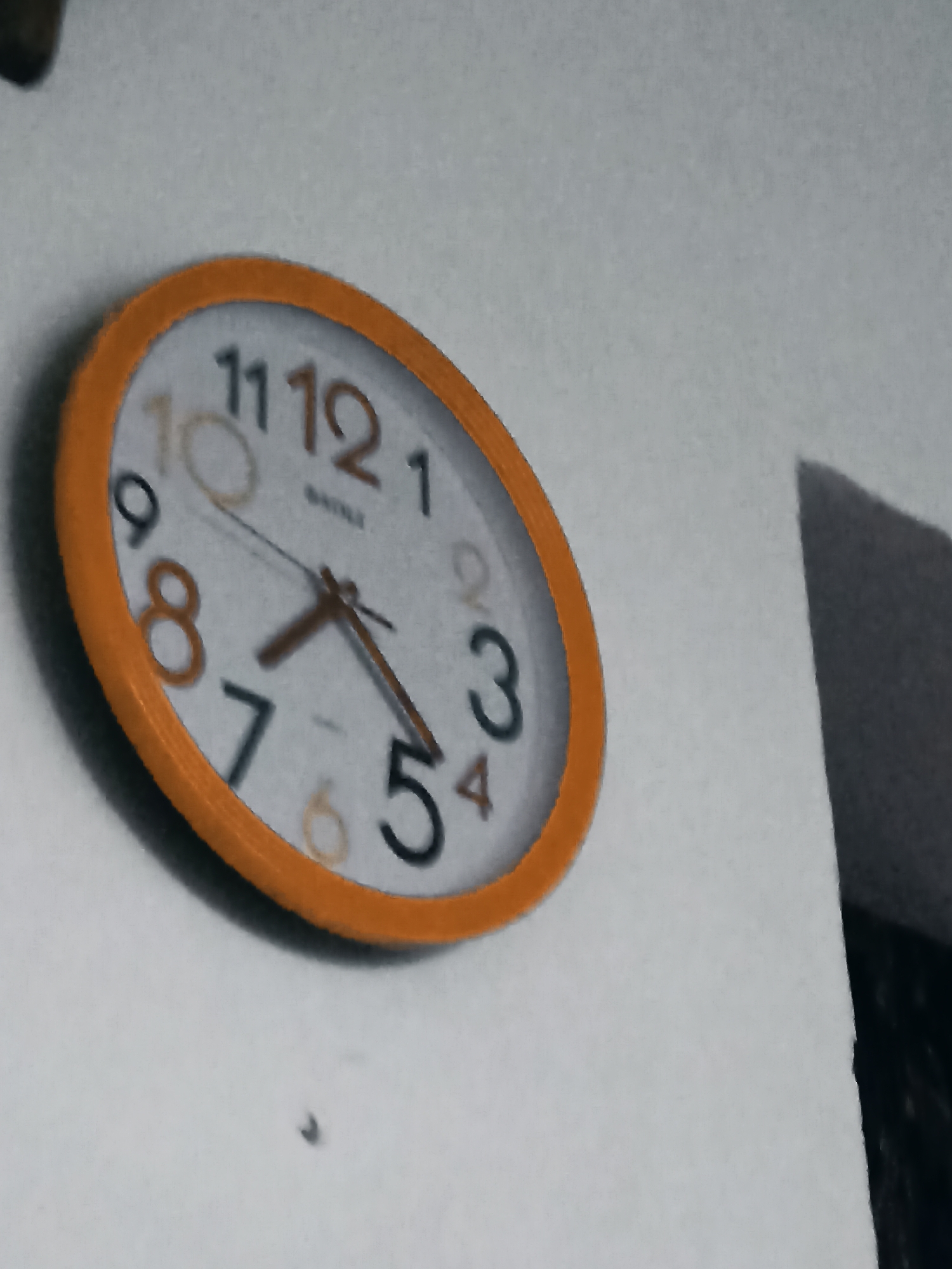 The image shows a close-up, slightly distorted view of an orange-rimmed analog clock mounted on a white wall. The clock face displays black Arabic numerals, with some of the numbers appearing slightly faded or a different color, possibly due to lighting or age. The hour hand is pointing between the 7 and the 8, appearing closer to the 7. The minute hand is pointing directly at the 5. The second hand is not clearly visible. The clock's hands are a dark brown color. The background is a plain, textured white wall. To the right of the clock, a dark, angled shadow is cast on the wall, suggesting a light source is present but not within the frame. There is a small dark speck on the wall below the clock. No people or any other objects are visible in the frame. The image quality is grainy, indicating it was taken with a low-resolution camera or in poor lighting conditions. The scene is indoors. There is no information or visual cues to suggest the location is in Ceyranbatan, Azerbaijan.