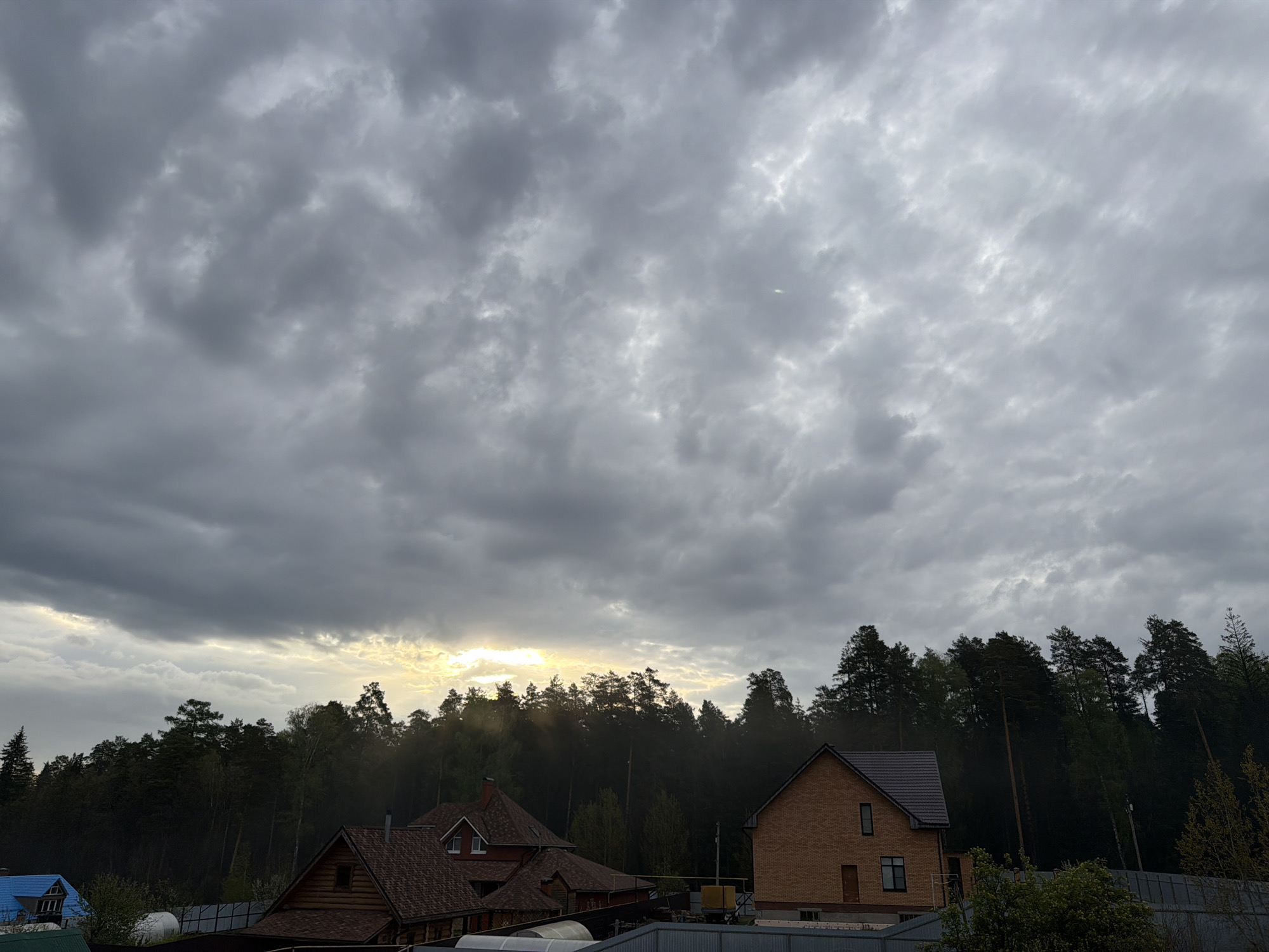 The image depicts a cloudy sky over a residential area in Chaikovsky, Russia. In the foreground, there are houses with tiled roofs and fences. The houses are of brick and wood construction. Behind the houses is a dense forest, with sunlight piercing through the trees. The sky is overcast with gray and white clouds.