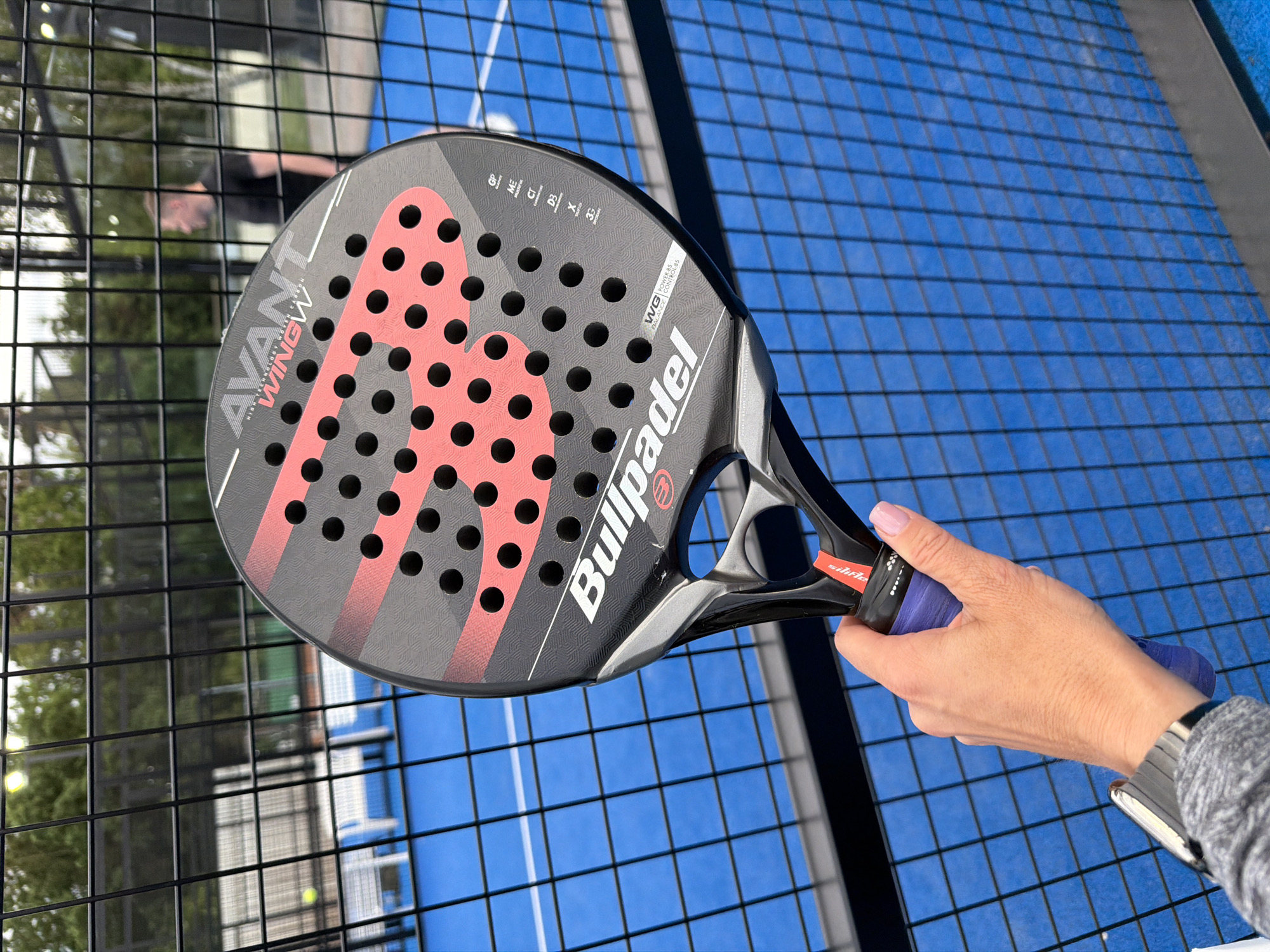 A person's hand, adorned with a smartwatch and pink-painted fingernails, holds a black and red "Bullpadel AVANT WINGW" padel racket in front of a blue padel court. The racket face features numerous black circular holes and a repeating pink patterned design. In the background, a black wire fence separates the court from a grassy area with trees. Beyond the fence, a person in a black t-shirt is visible walking away from the camera. Several white chairs are positioned on the court. The scene appears to be an outdoor recreational facility.