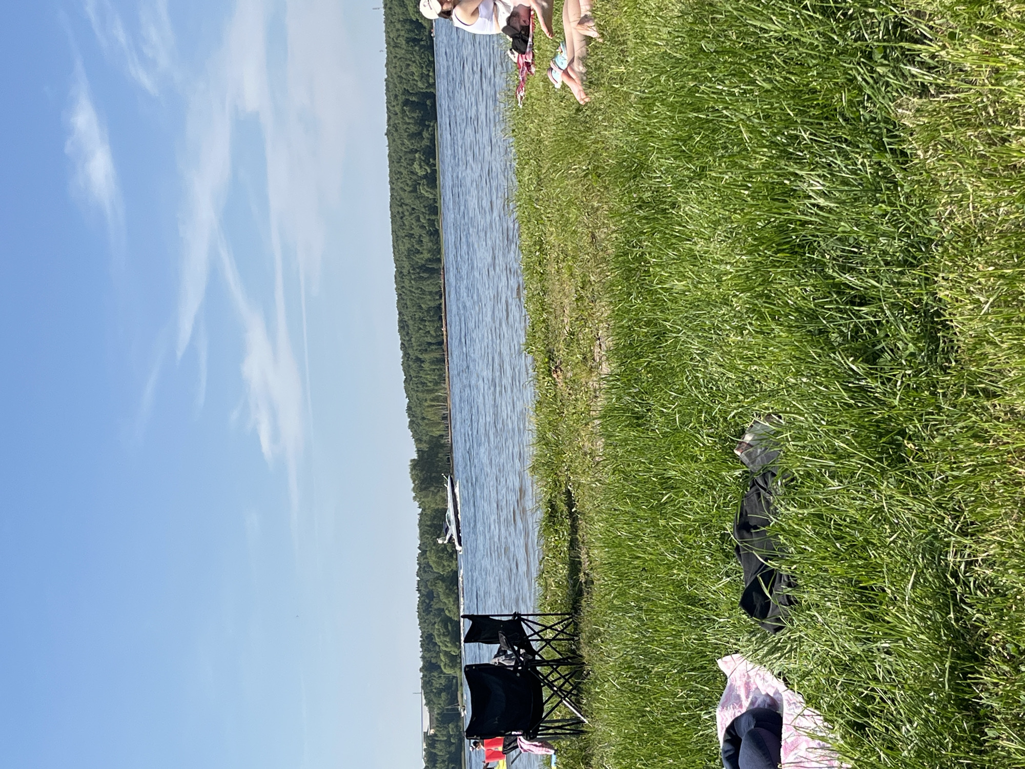 This image captures a wide-angle view of a recreational scene along the grassy bank of a body of water in Kapustino, Russia, under a clear, bright blue sky with scattered white cirrus clouds.
In the immediate foreground, lush green grass occupies the lower half of the frame. To the left, two black folding camping chairs are positioned on the grass. Adjacent to the chairs on the left, a pink and white patterned blanket or towel is spread on the ground, with a dark blue item (possibly clothing) partially visible on it. To the right of the chairs, a black piece of clothing or fabric lies on the grass.
The midground features the water body, which appears calm with minor surface ripples. A dark-colored motorboat, generating a distinct wake, traverses the water from left to right. Further to the left on the water, a small white sailboat is faintly visible in the distance. Along the grassy bank on the right side of the frame, multiple individuals are present. One person, wearing a white tank top and a light-colored baseball cap, sits upright with their back partially turned towards the camera. Below and to the right of this individual, two sets of bare legs are visible, indicating at least two other people are lying down on the grass, largely obscured by vegetation. On the left side of the bank, near the black chairs, another person is partially visible, wearing a red and yellow life vest, facing away from the camera.
The background consists of a dense, dark green forest line forming the opposing shoreline of the water body. Beyond the trees on the far left horizon, faint industrial or building structures are discernible. The overall lighting suggests midday conditions.