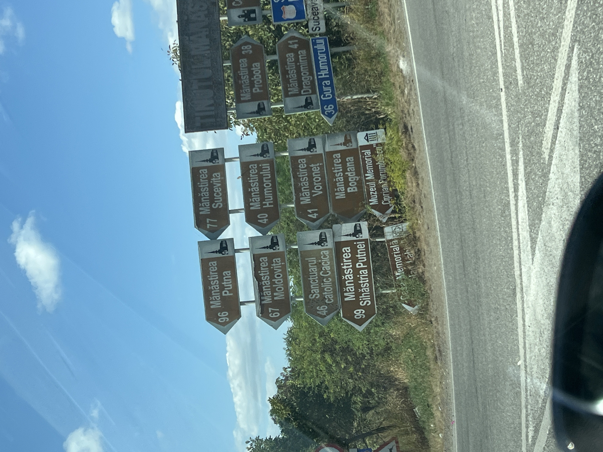 The image captures a roadside scene with multiple directional signs pointing towards monasteries and other points of interest in the region of Cotu Băii, Romania. The signs, predominantly brown with white lettering and symbols of churches, indicate distances in kilometers and names of locations such as "Mănăstirea Putna," "Mănăstirea Sucevita," "Mănăstirea Moldovita," "Sanctuarul catholic Cacica," "Mănăstirea Sihastria Putnei," "Mănăstirea Humorului," "Mănăstirea Voronet," "Mănăstirea Probota," "Mănăstirea Dragomirna," "Mănăstirea Bogdana," "Gura Humorului," and "Muzeul Memorial Ciprian Porumbescu." The signs are mounted on poles, with some visible in the foreground and others further in the background. In the upper right corner, a larger dark sign with light lettering appears to read "TINUTUL MANAS." The background consists of lush green trees and foliage under a blue sky with scattered white clouds. A paved road with white lane markings is visible in the foreground, with the edge of a vehicle's windshield in the lower portion of the frame, suggesting the photograph was taken from inside a car. The environment appears to be rural or semi-rural. No people are visible in the image.