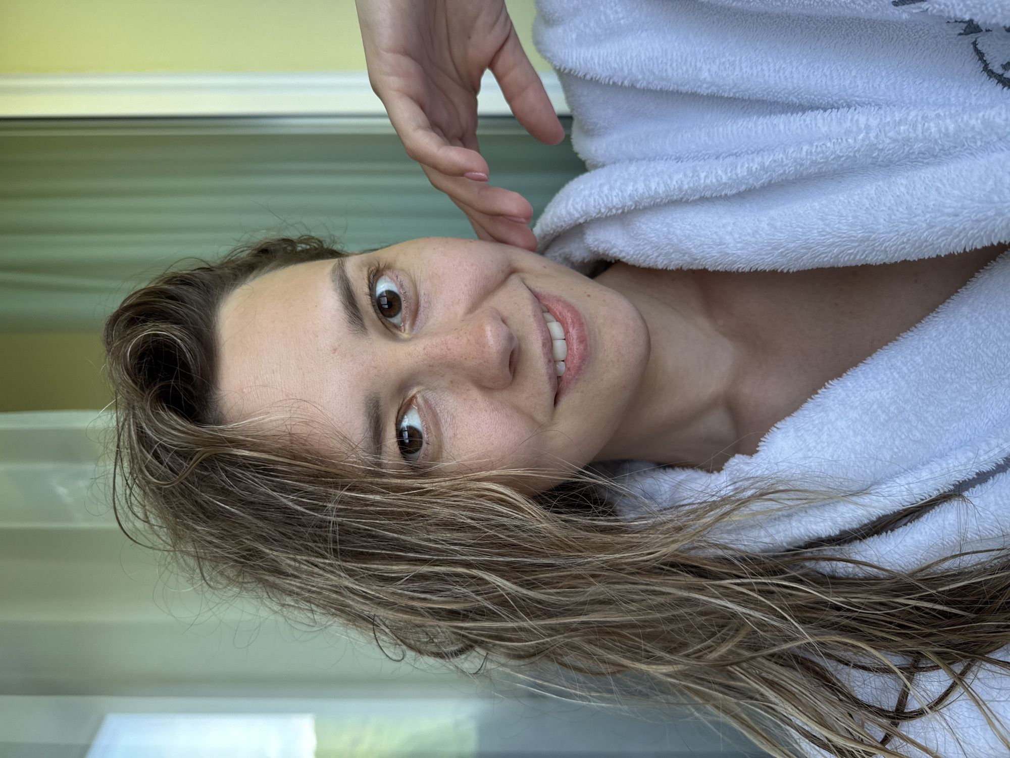 A frontal, chest-up portrait of a female individual with damp, disheveled light brown hair with blonde highlights, falling over her shoulders and partially obscuring the left side of her face. She has brown eyes, visible freckles across the nose and cheeks, and a slight, closed-mouth smile. She is wearing a white, plush terrycloth bathrobe. Her left hand is positioned with fingers gently touching her left jawline/cheek area. The background consists of an indoor setting with a large window or sliding glass door on the left, revealing a bright sky, and a light green wall or panel on the right. The scene is illuminated by natural light. The location is Nebug, Russia.