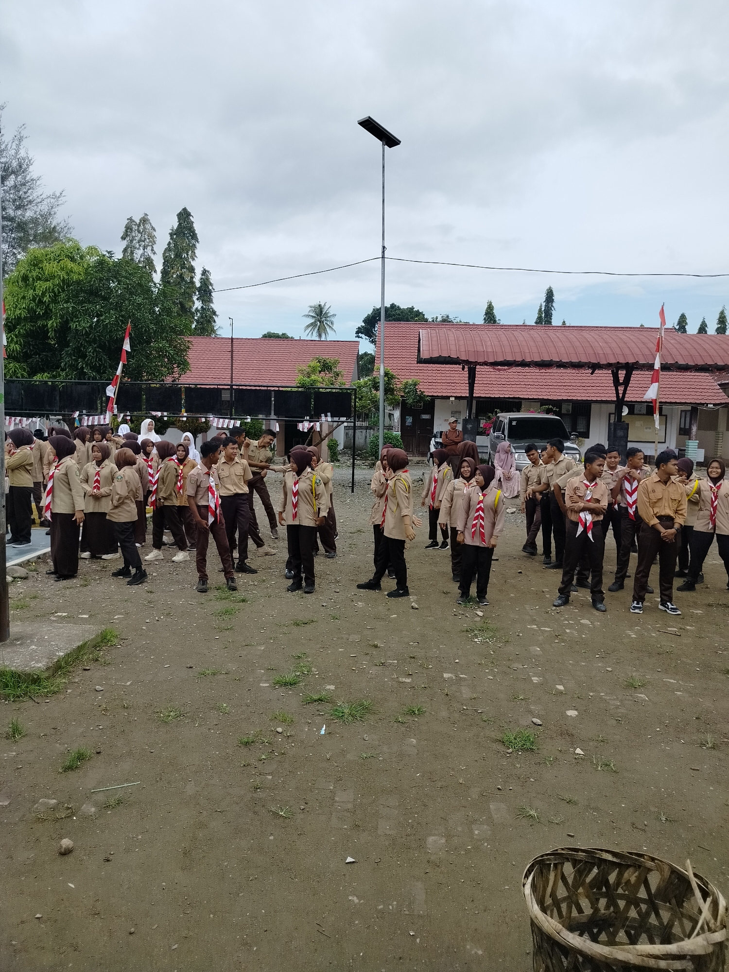 The image depicts an outdoor scene in Purwodadi, Indonesia, featuring a group of people, predominantly young individuals dressed in scout uniforms, gathered in an open area. The setting appears to be school grounds or a similar communal space, with buildings featuring red-tiled roofs visible in the background.
The individuals are standing in various formations, some engaged in conversation or seemingly waiting for instructions. Their attire consists of khaki-colored shirts and brown trousers or skirts, adorned with red and white scout neckerchiefs. A few adults are also present, some observing the group.
The environment is characterized by a dirt and gravel ground, with patches of grass growing sporadically. A lamppost with a solar panel is prominently positioned in the middle ground, and a woven basket is visible in the foreground. Indonesian flags, red and white, are displayed on flagpoles and attached to some of the buildings, suggesting a national event or observance. The sky is overcast, indicating a cloudy day. The overall atmosphere suggests an organized gathering or activity, possibly a scouting event or a school assembly.