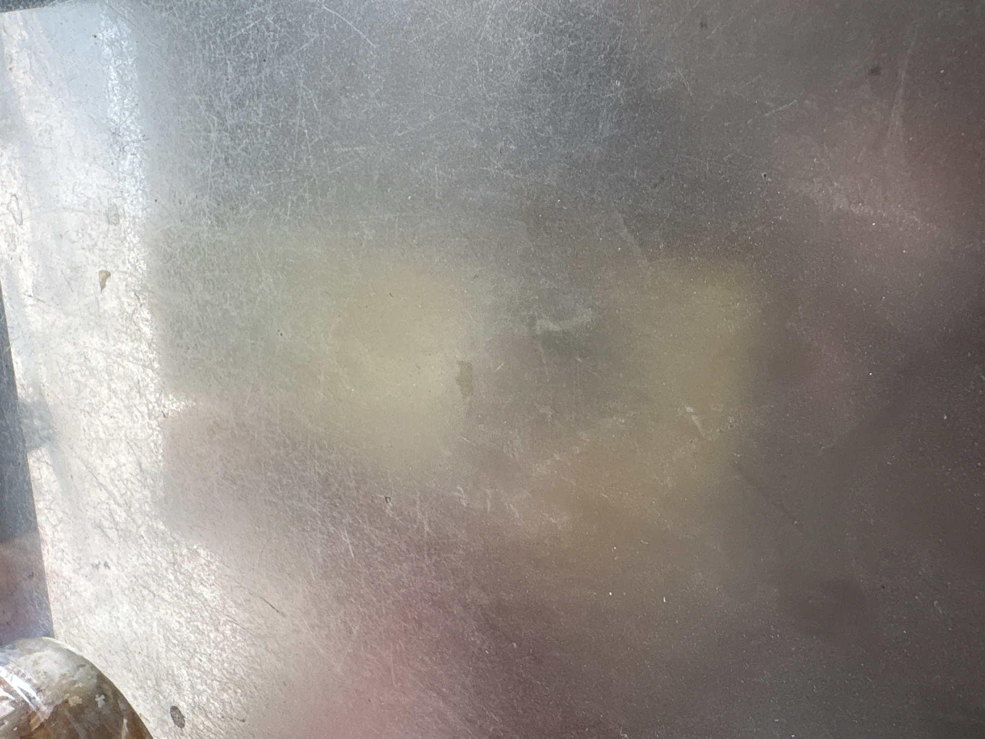 The image shows a close-up of a scuffed metal surface, likely a tabletop, with visible scratches and smudges. A blurred, rounded object with a textured surface, possibly a jar or container, is partially visible in the upper left corner. The lighting creates diffuse reflections and highlights on the metal. There are no discernible people or specific landmarks that would indicate a location in Palimanan, Indonesia. The scene appears to be an indoor setting, given the lighting and the material of the surface.