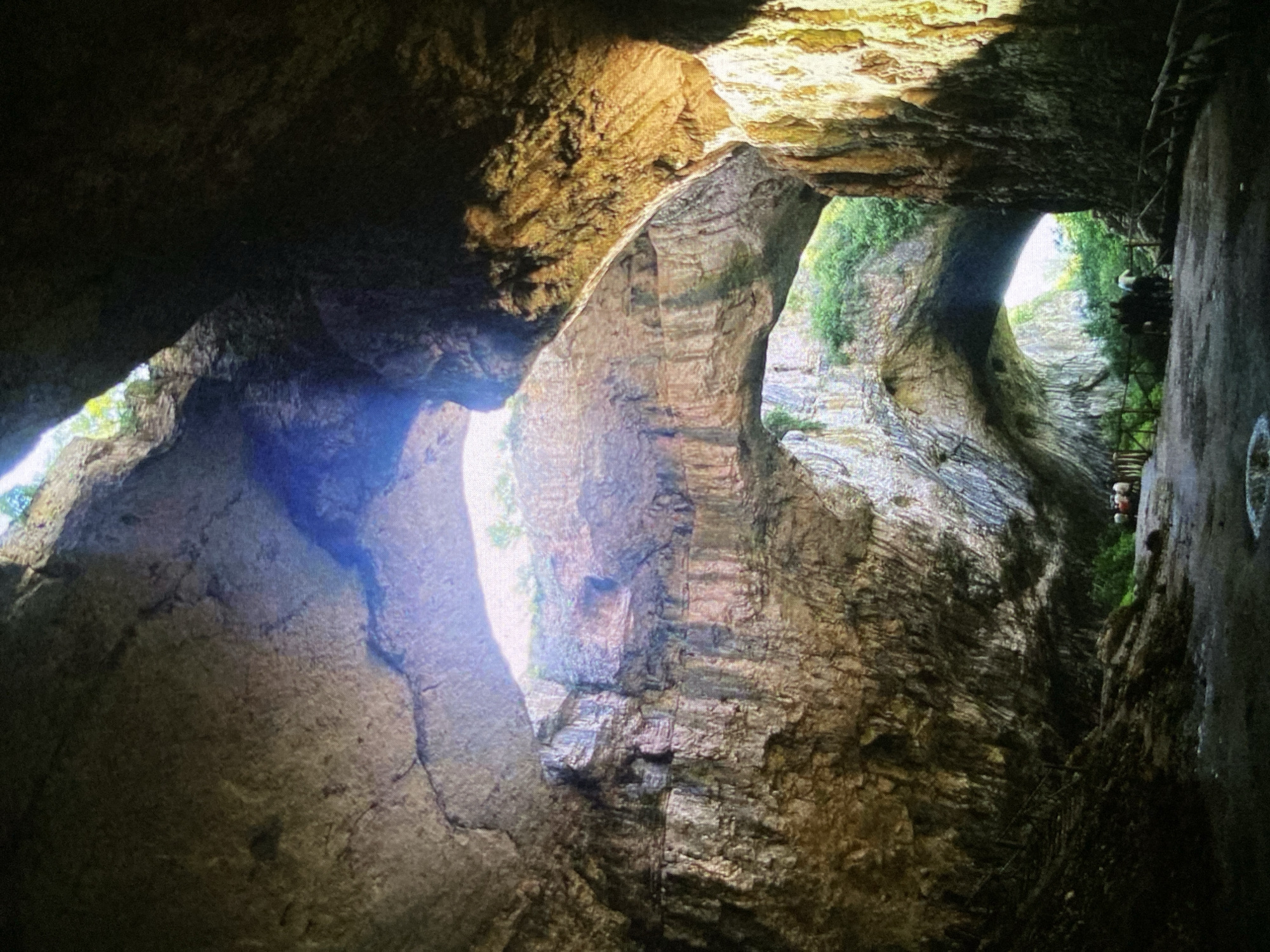 The image captures a dramatic view inside a deep natural gorge or canyon, possibly a cave entrance, characterized by massive rock formations. Strong sunlight penetrates from openings above and through archways, creating stark contrasts and highlighting the rugged, textured surfaces of the rock walls. These walls exhibit visible stratification and varying shades of brown, gray, and green, indicative of natural geological processes.
Along the right side of the chasm, a wooden walkway or staircase is constructed into the rock face, providing access for visitors. Two individuals are clearly visible on this suspended structure, suggesting they are exploring or observing the natural surroundings. Hints of green vegetation, possibly moss or small plants, cling to the rock where light reaches, and more abundant foliage is seen through some of the openings.
The scene appears to be set during the daytime, with the bright, direct sunlight indicating clear weather. The overall impression is one of natural grandeur and scale, with the human figures providing a sense of perspective within the immense rock formations. The image does not contain any visible text. Based on the context, this location is a natural attraction in Bankya, Bulgaria, designed for visitors to experience its unique geological features.