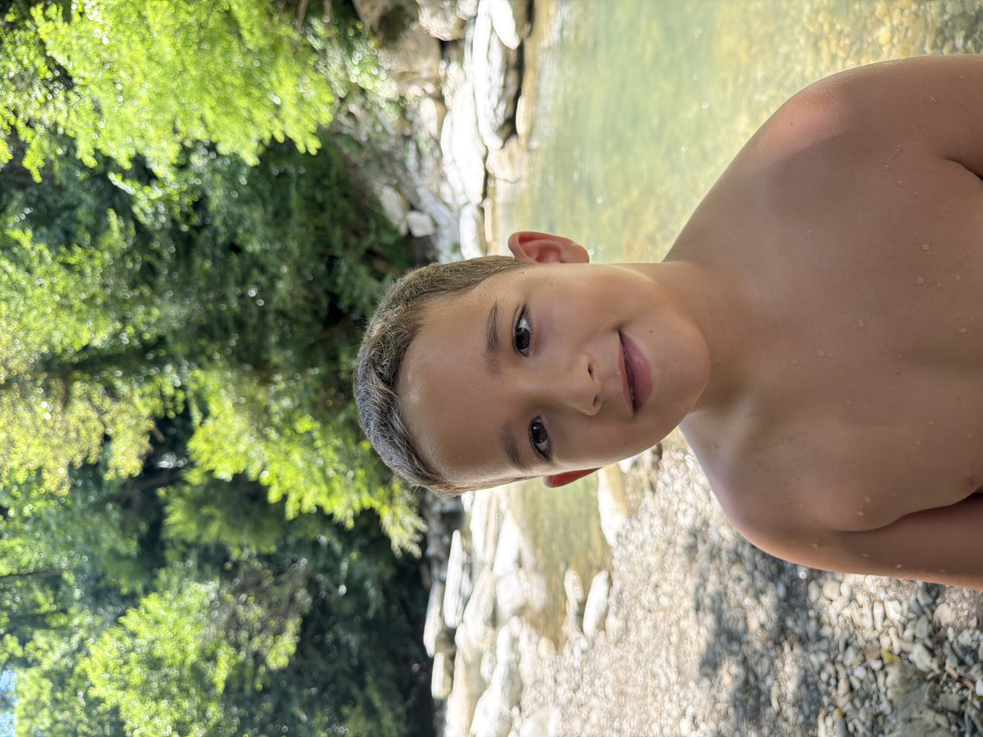 The media file shows a close-up, angled portrait of a young boy. He is positioned in the foreground, looking towards the camera with a slight smile. His skin is wet, suggesting he has recently been in water. The background features a body of clear, shallow water with visible rocks and pebbles at the bottom, and a rocky bank lined with lush green trees and foliage. The lighting indicates it is daytime, likely with bright sunlight filtering through the trees, casting dappled shadows. The overall setting appears to be a natural, outdoor environment, possibly a river or stream in a wooded area. The boy's expression is relaxed and happy, conveying a sense of enjoyment in his surroundings. There is no visible text in the image.