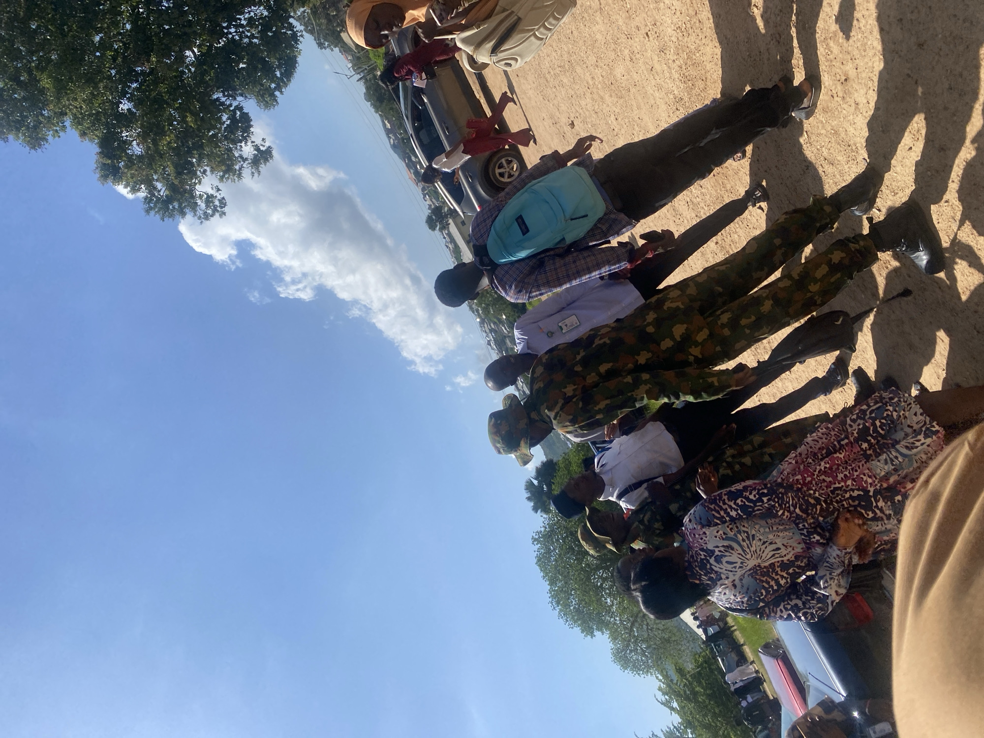 In Jos, Nigeria, under a bright blue sky with scattered white clouds, a group of people are gathered on a dirt path. The scene appears to be outdoors in a semi-urban or suburban area, with some trees and buildings visible in the background.
Several individuals are dressed in military camouflage uniforms, including one person in the foreground who is the primary subject, facing away from the camera. This person is wearing a camouflage jacket and trousers, with a boonie hat. Another individual in camouflage is also visible in the mid-ground.
Interacting with the military personnel are civilians. One woman in a brightly colored patterned dress is looking towards the group. Another person, a man in a plaid shirt and jeans, is seen with a blue backpack. Other individuals in civilian attire, some appearing to be students in white shirts, are also part of the gathering.
In the background, several vehicles are parked, including what appears to be a silver SUV and another dark-colored car. The environment is dusty, with shadows cast on the ground by the people and objects, indicating it's a sunny day. The overall impression is a casual gathering or interaction, possibly at a public space or event in Jos.