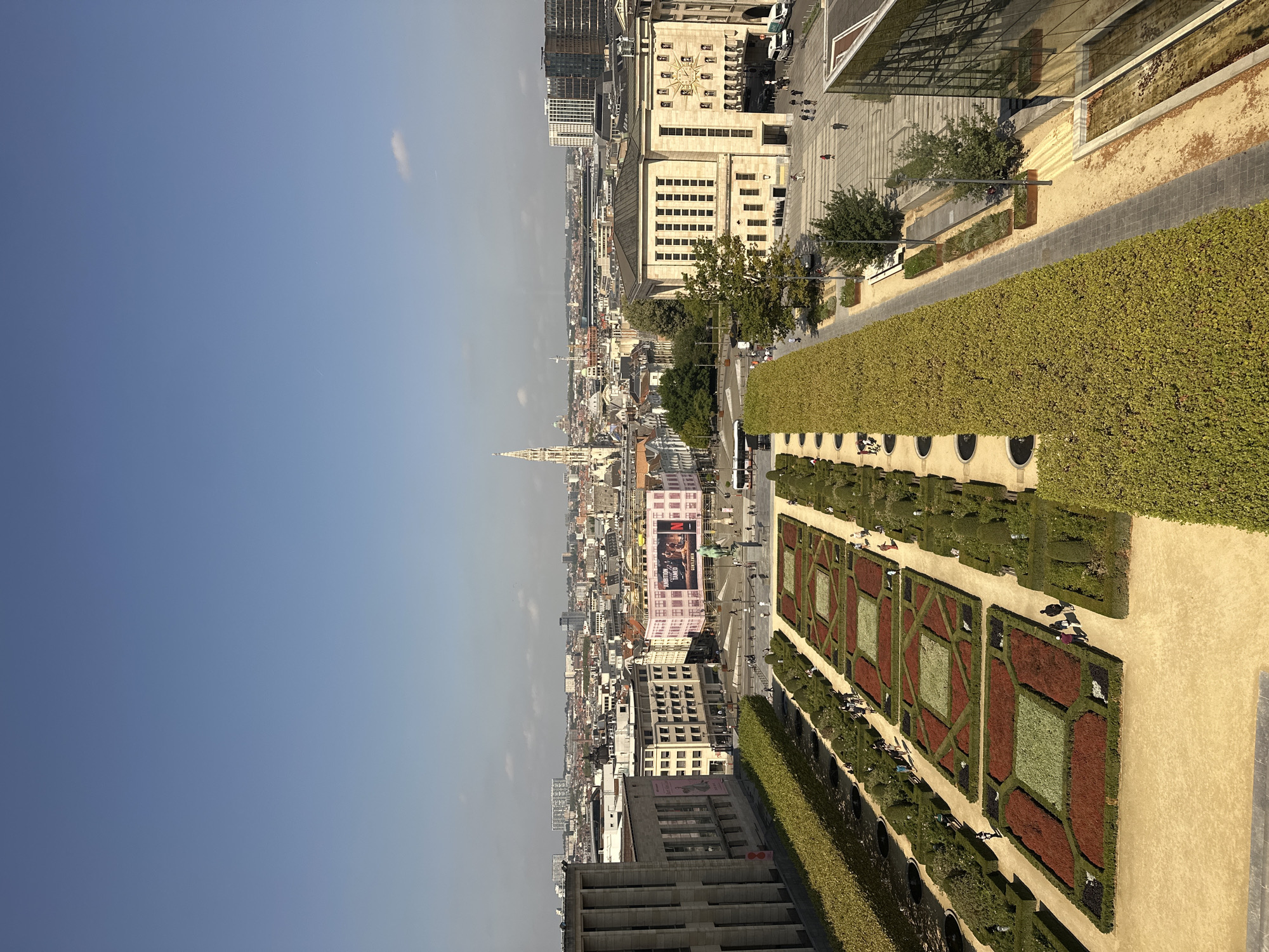 This is a high-angle, eye-level, full shot of the Mont des Arts in Brussels, Belgium. The foreground features a meticulously manicured French formal garden with geometric flower beds filled with red, green, and dark foliage. The garden is bordered by manicured hedges and pathways. People are observed walking through the garden and along the pathways.
In the mid-ground, there is a wide plaza with a large building on the right that has a prominent clock face on its facade. This building appears to be the Town Hall of Brussels. Across the plaza are several other buildings, including a large pink building with billboards displaying advertisements for "AMBITION TAMED" and "GENTLEMEN". A statue of a man on a horse is visible in front of the pink building. Buses and cars are present in the plaza.
The background reveals the sprawling cityscape of Brussels, characterized by a mix of historical and modern architecture. A tall spire, likely part of the Cathedral of St. Michael and St. Gudula, stands out against the skyline. The sky is clear and blue, indicating a sunny day. The overall scene depicts a vibrant urban landscape with historical landmarks, public spaces, and a well-maintained garden.