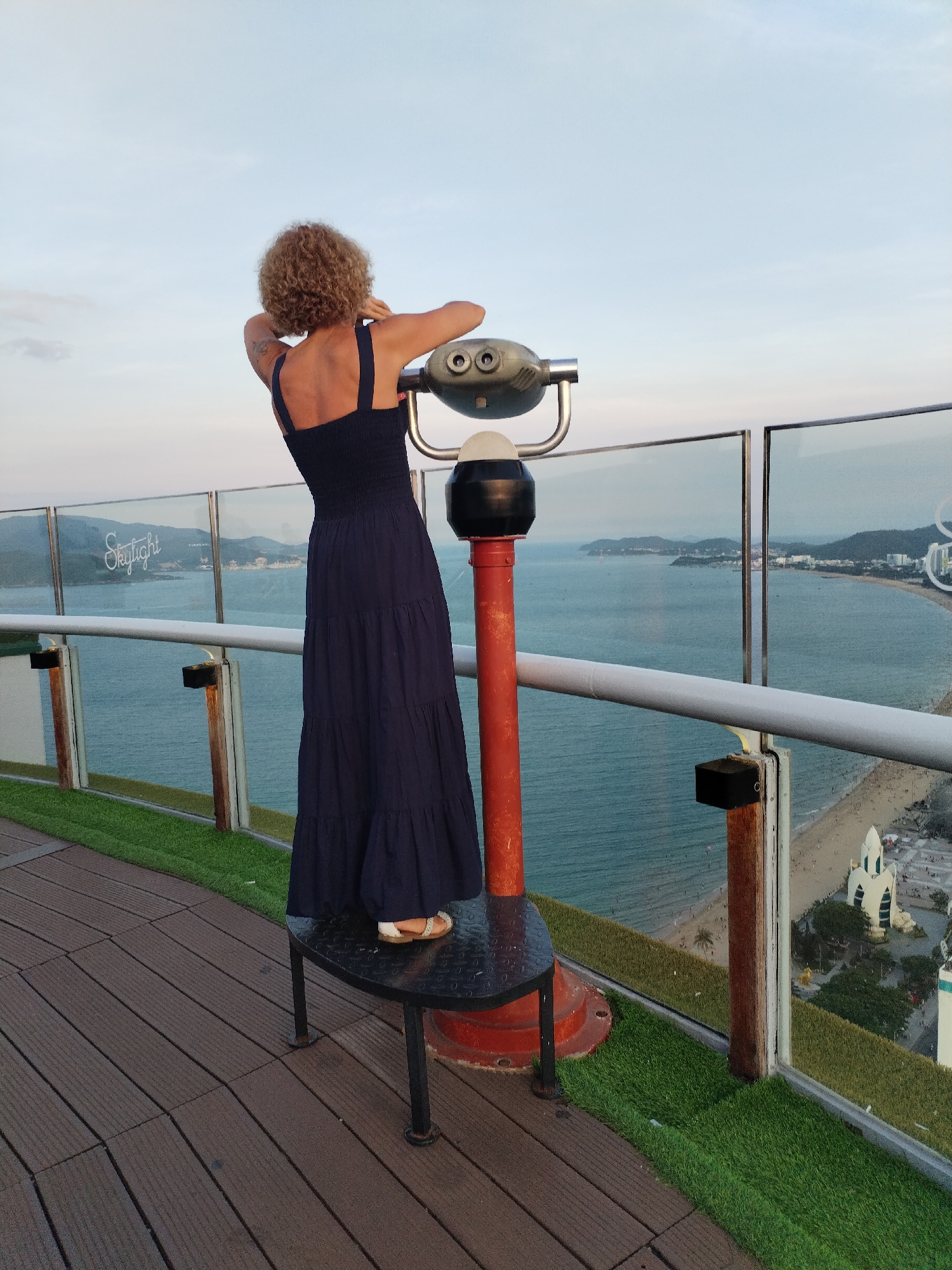 An individual with curly blonde hair, wearing a dark blue, tiered maxi dress and sandals, stands on a dark metal platform. They are positioned with their back to the camera, looking through a coin-operated telescope. The telescope is mounted on a red pole. The scene is set on a wooden deck with artificial turf bordering a glass railing. Beyond the railing, a panoramic view unfolds: a bay with calm turquoise water, a sandy beach populated with numerous people, and a cityscape with buildings nestled against rolling hills in the distance. The sky is a pale blue with hints of clouds, suggesting it is either early morning or late evening. The location is identified as Nha Trang Ward, Vietnam.