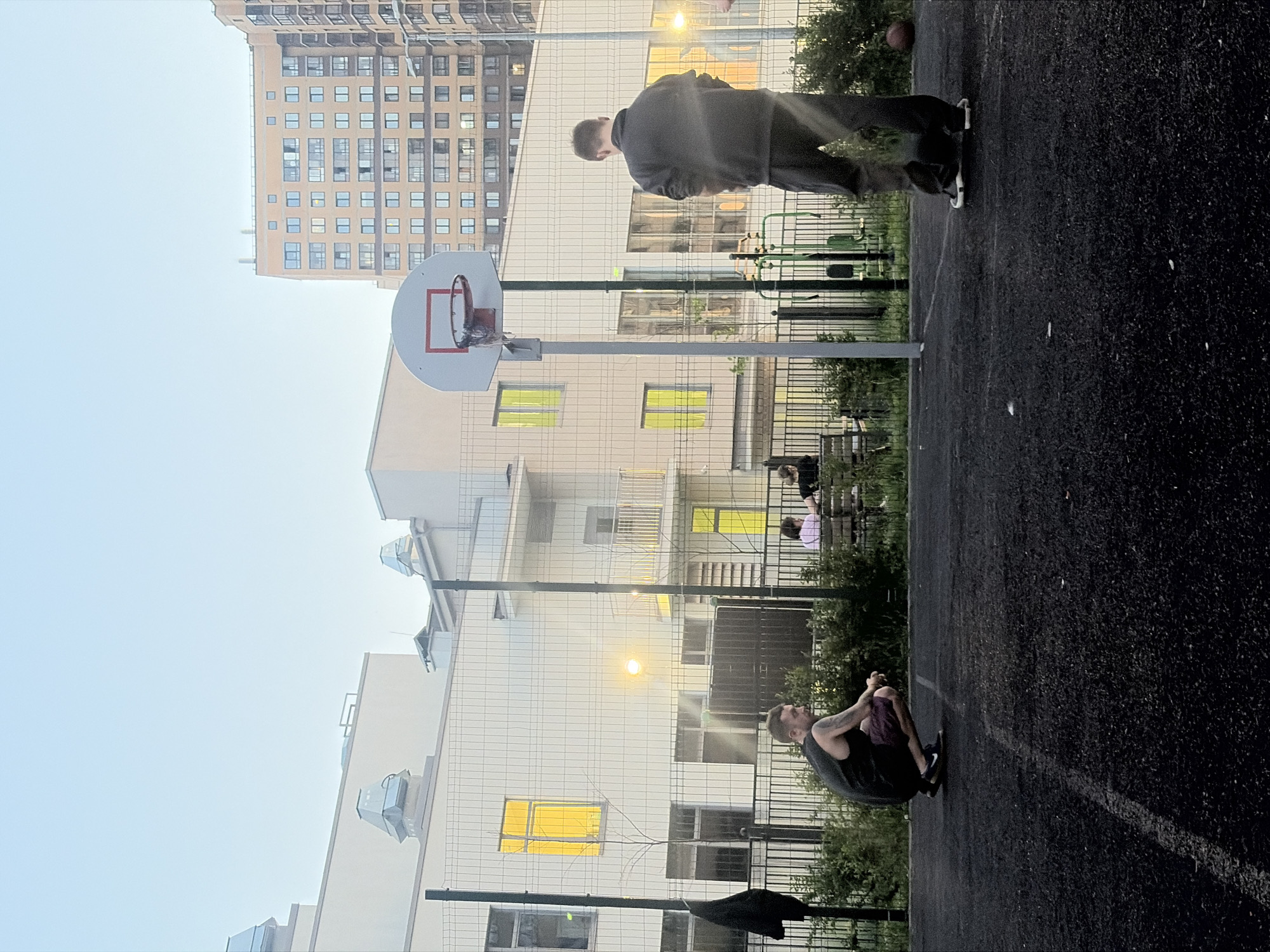 This photo appears to depict a basketball court located in Saint Petersburg, Russia. A man in a grey jacket is standing by the basketball hoop. Another man is sitting on the ground near the court, seemingly stretching. In the background, there is a tall apartment building. The day appears to be overcast. There is a fence separating the court from the surrounding area. Other people are sitting on a bench in the background.