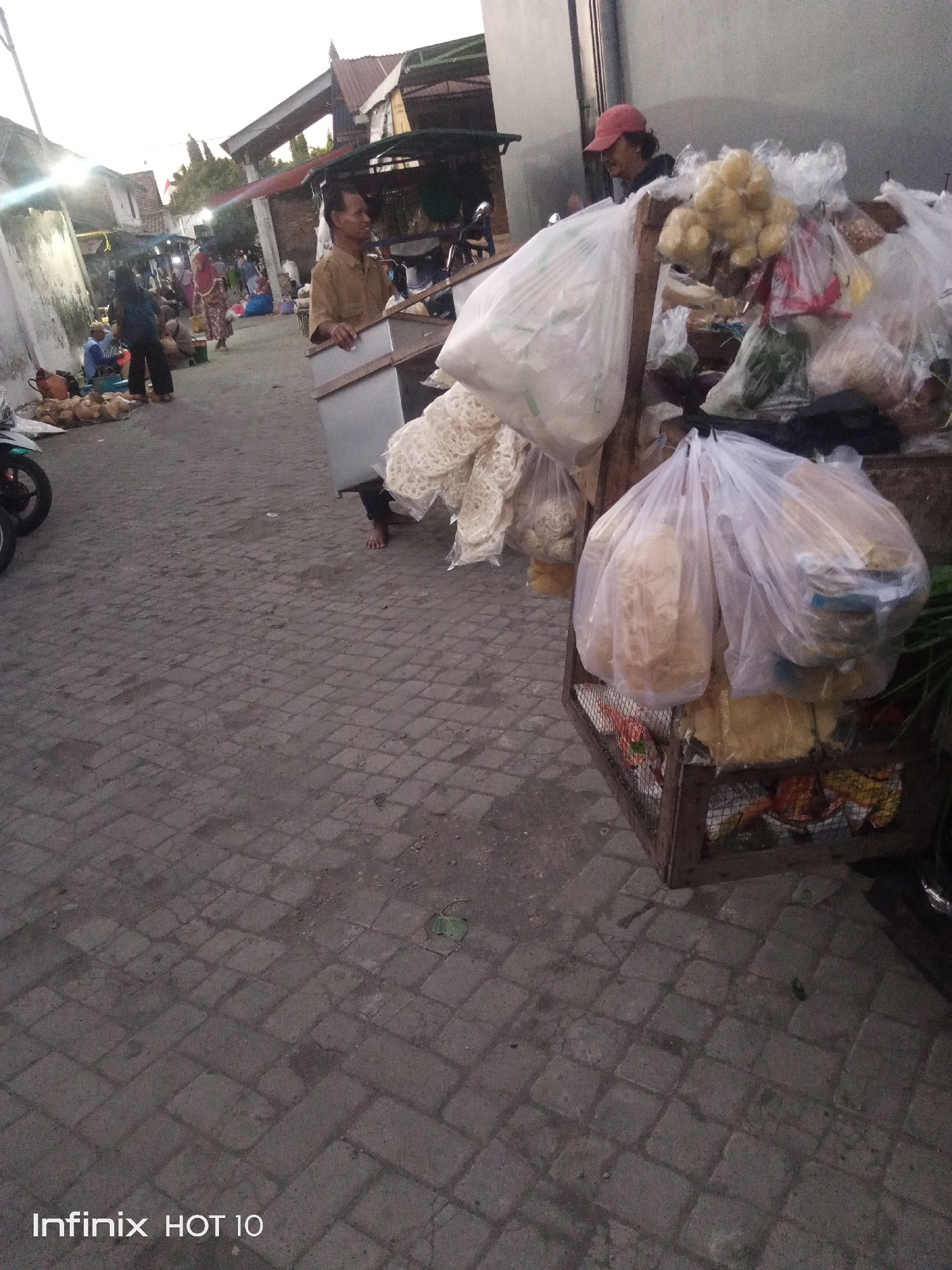 The image depicts an outdoor scene in the city of Sono Ageng, Indonesia, likely a market or street-side vendor area. A man wearing a tan shirt is visible, carrying a metal container. He appears to be a vendor, as he is standing next to a stall filled with various food items packaged in plastic bags, including what appears to be crackers or puffed snacks. Another person, wearing a red cap, is partially visible in the background, also seemingly involved with the vendor stall. In the background, other individuals and market stalls are present, suggesting a bustling commercial environment. The ground is paved with interlocking bricks. The overall atmosphere suggests an everyday commercial activity taking place in a moderately populated urban setting. The "Infinix HOT 10" watermark at the bottom indicates the device used for capture.