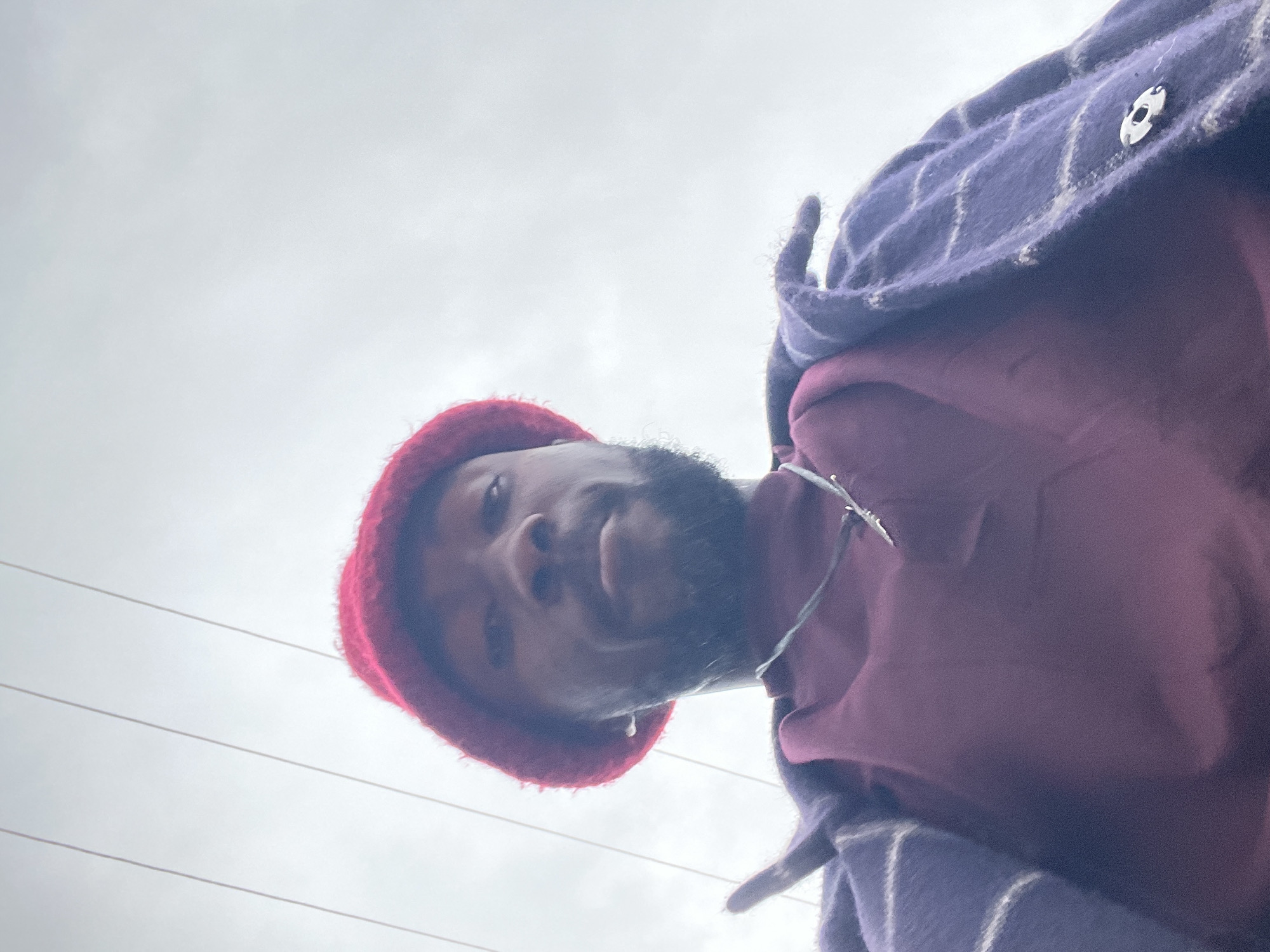 A low-angle, close-up selfie shows a Black man wearing a red knit hat and a purple and white plaid jacket over a maroon turtleneck. He has a beard and is looking directly at the camera. The background is a gray, overcast sky with power lines visible across the top. The scene appears to be outdoors in an urban environment, possibly Jos, Nigeria, given the overcast sky and visible power lines, which are common features in such settings. The man is centered in the frame, with his face and upper body dominating the image.