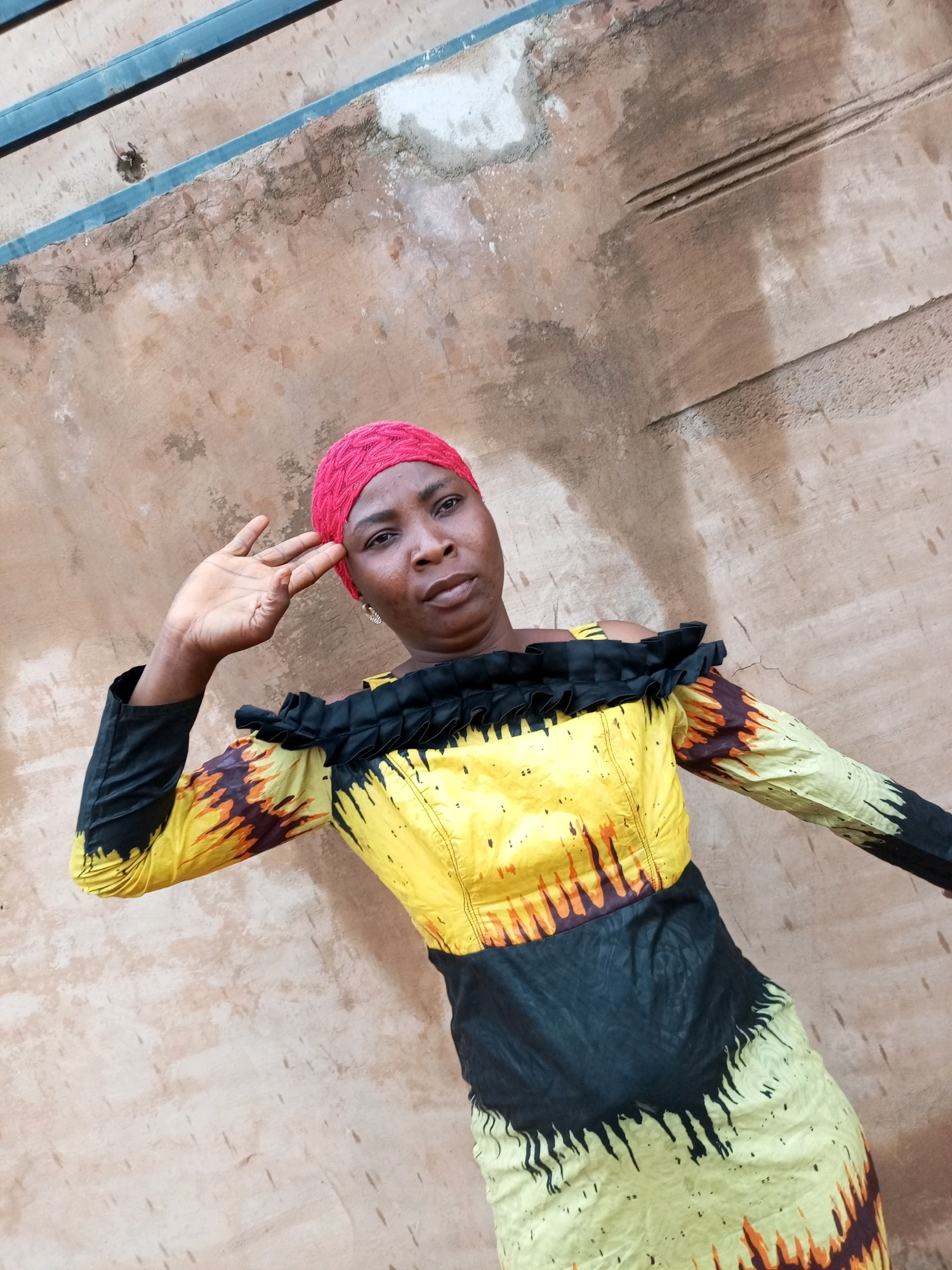 A woman wearing a bright pink headscarf and a yellow and black off-the-shoulder dress is posing against a textured, beige-colored wall in Tempure, Nigeria. The dress features a ruffled black neckline and a vibrant yellow and black pattern on the sleeves and skirt, with orange accents. The woman's right hand is raised to her forehead, with her fingers extended. The wall behind her appears weathered and has some faint lines or markings. Above the wall, a blue metallic structure is visible, likely a part of a building or fence. The lighting suggests it is daytime.