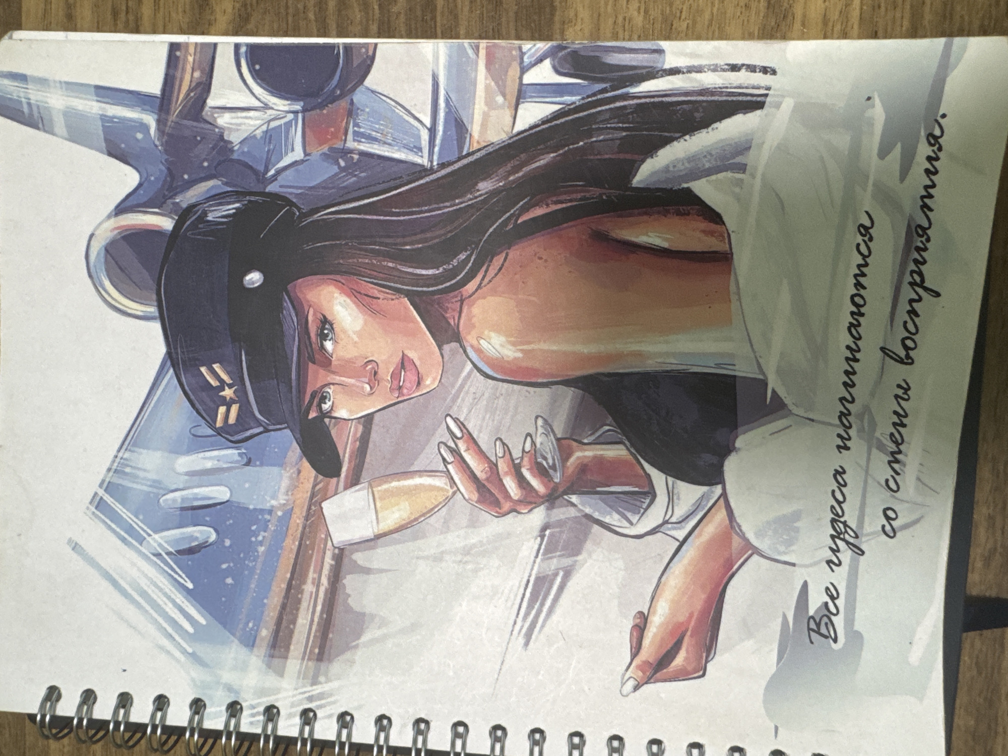 A spiral-bound notebook lies on a wooden surface. The notebook cover features a stylized illustration of a woman in a pilot's hat holding a glass of champagne. She is positioned in front of an abstract depiction of an airplane. Below the illustration, in cursive script, are the words "Все чудеса начинаются со смены восприятия" which translates to "All miracles begin with a change of perception."