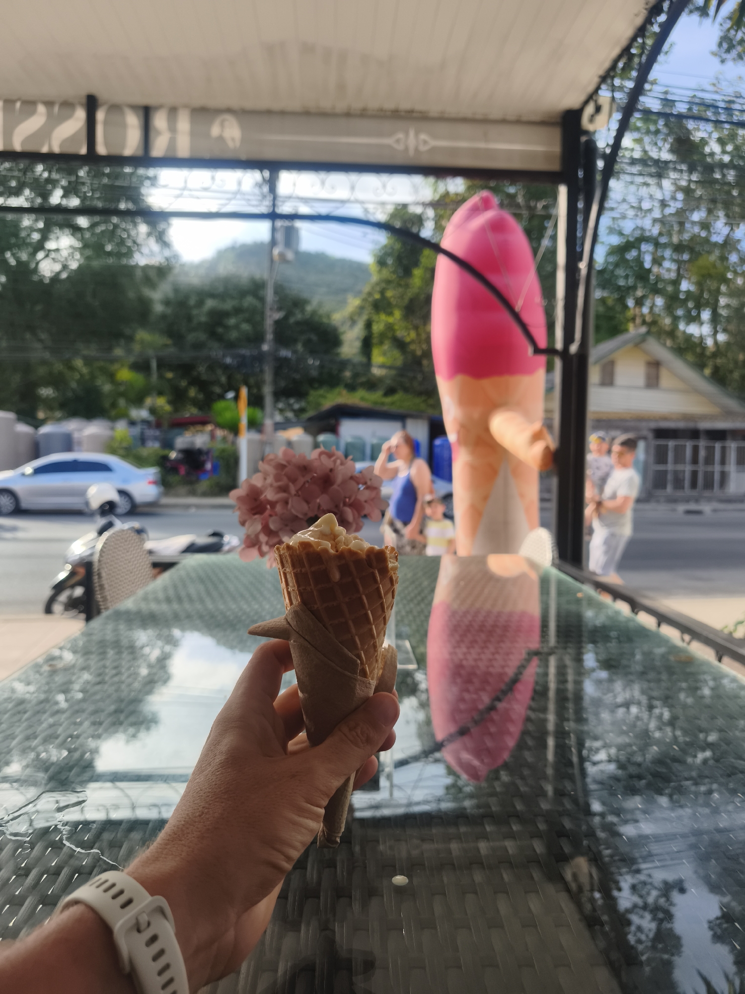 A person holds an ice cream cone in their hand, seemingly indoors at a cafe or shop. The ice cream cone is a waffle cone with what appears to be vanilla or caramel ice cream on top. The person is wearing a white smartwatch. The scene is viewed through a window, showing a street scene in Baan Chaweng Noi, Thailand.
Visible outside are people, a car, a motorbike, and a large inflatable ice cream cone used for advertising. A building and foliage can be seen across the street. The location appears to be a cafe or shop where customers can sit, based on the glass-topped table in the foreground.