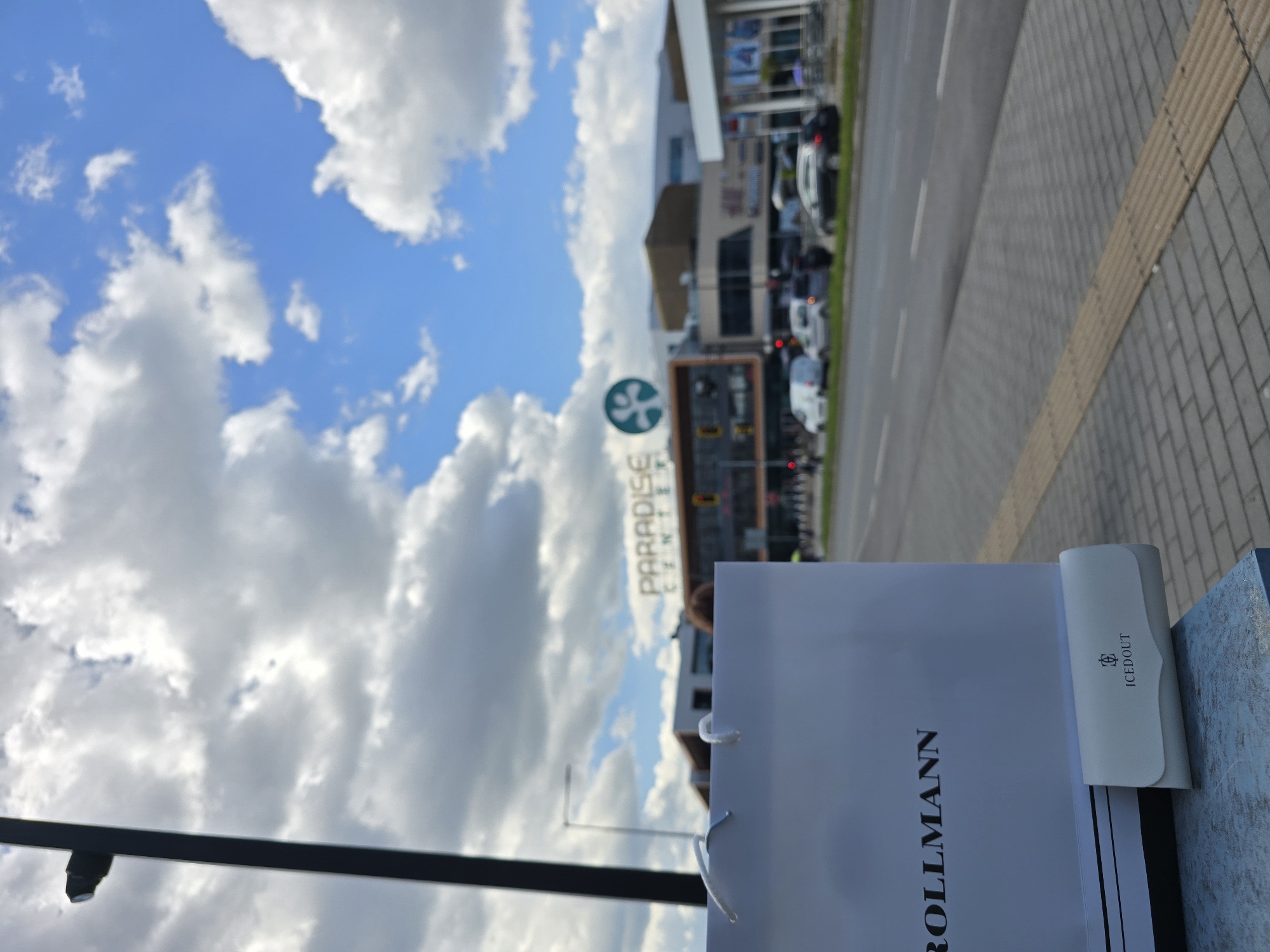 The image shows a daytime view in Sofia, Bulgaria. A shopping bag with the word "ROLLMANN" is prominently displayed in the foreground, with a white ICEDOUT case nearby. In the background, the "PARADISE CENTER" sign is visible above a building, which is likely the Paradise Center shopping mall. The sky is blue with fluffy white clouds. A street and sidewalk are also visible, with cars and other vehicles driving on the road.