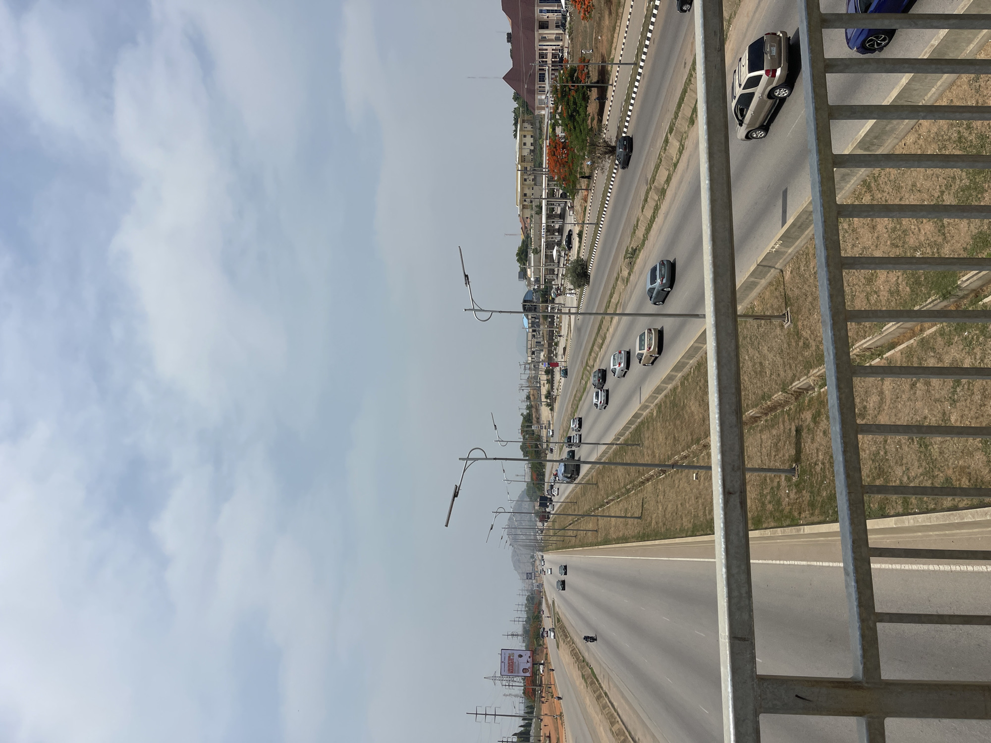 The image depicts a multilane highway in Kuchingoro, Nigeria, viewed from an elevated position, likely a pedestrian bridge. The sky is overcast with scattered clouds. Two sets of lanes run parallel, divided by a median. Cars are visible in both directions. Streetlights line the highway. Buildings and vegetation are visible on the far side of the highway. A metal railing is in the foreground.
