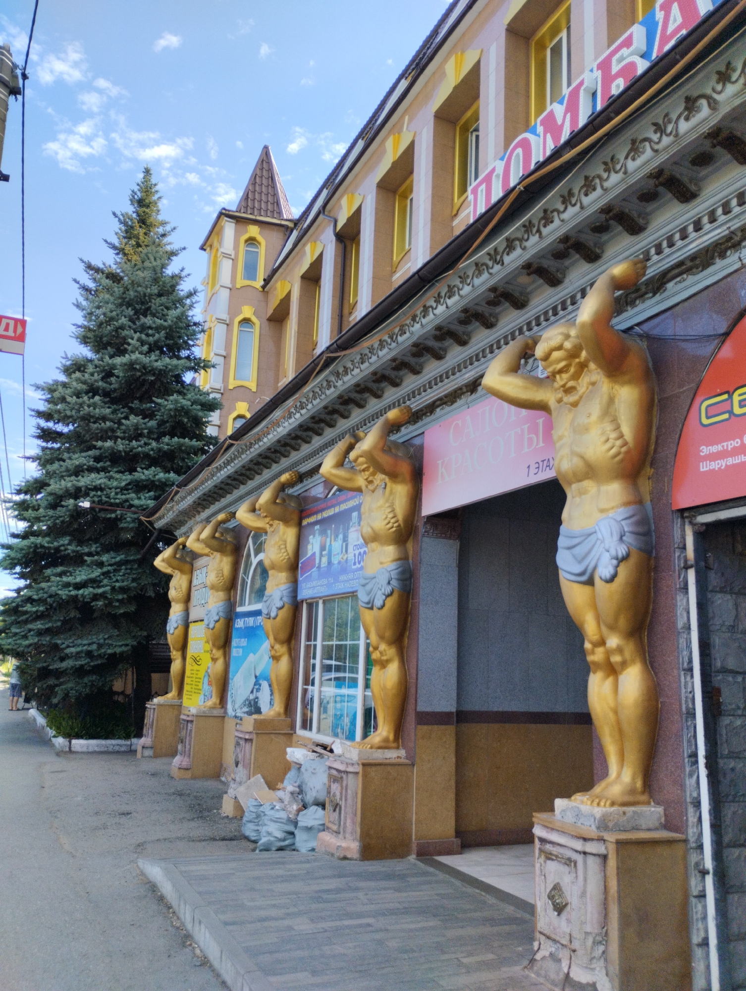 The image shows a street view of a building with a prominent gold-colored facade, featuring several statues of men holding up the building's overhang. The building has a reddish-brown and yellow color scheme with ornate architectural details. Large, green coniferous trees are visible on the left side of the image, and a blue sky with scattered clouds is overhead. The street in front of the building is paved with grey and brown tiles, and there are some trash bags and debris on the sidewalk. Text in Russian is visible on the building, including what appears to be "САЛОН КРАСОТЫ" (Beauty Salon) and other signage. The overall scene suggests an urban environment in a location with warm weather, given the clear sky and light clothing of a person partially visible on the far left. Based on the architectural style and the visible Russian text, this building is likely located in a city with a significant Russian-speaking population. Without additional information or context, it is impossible to definitively confirm the location as Kostanay, Kazakhstan.