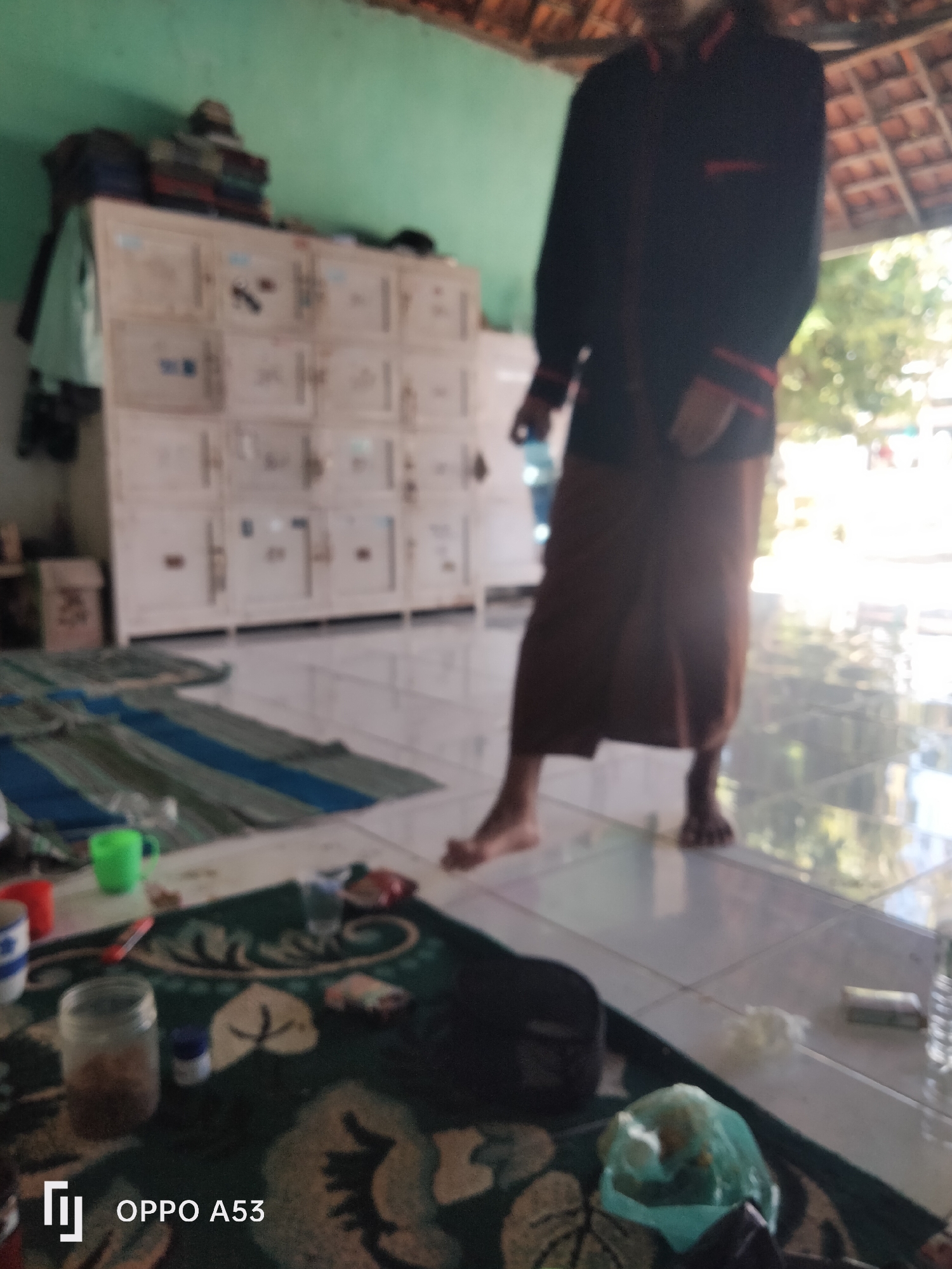 A person wearing a dark jacket with red trim and a brown sarong stands on a tiled floor, partially obscured by a light blue plastic bag containing food and a black bag. In the foreground, a patterned rug features stylized leaf motifs. To the left, a glass jar with a brown substance and a small blue container are visible, along with a green mug and a red mug. In the background, a row of white lockers with individual doors and handles is stacked against a light green wall. Above the lockers, stacks of fabric items are present. A thatched roof structure extends from the right side of the frame, allowing sunlight to illuminate the scene. The floor is tiled with white square tiles that reflect the light, and striped mats are laid out on the floor in the midground. The scene appears to be indoors, with an open-air area or courtyard visible through the roof. The overall image is blurry, suggesting it was taken with a mobile phone. The bottom left corner of the image displays the "OPPO A53" logo.