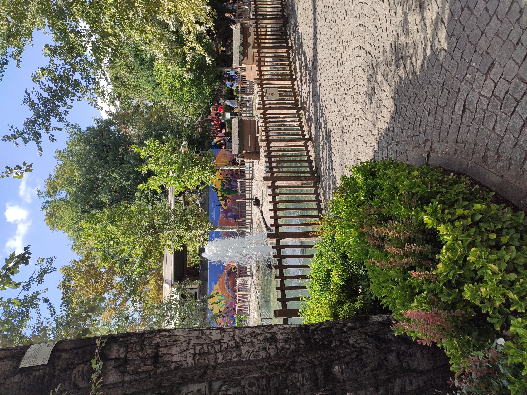 The image depicts a park scene in Ankara, Turkey, featuring a fountain and a pond surrounded by wooden fencing. Lush green bushes and trees frame the area, with a cobblestone pathway in the foreground casting shadows from the sunlight filtering through the leaves. In the background, a colorful mural adorns a wall, and several people are visible, some standing near a structure that appears to be a cafe or restaurant. There are also two duck-like figures near the pond and some wooden structures resembling animal shelters. The overall atmosphere is serene and inviting, typical of a public park in a city.