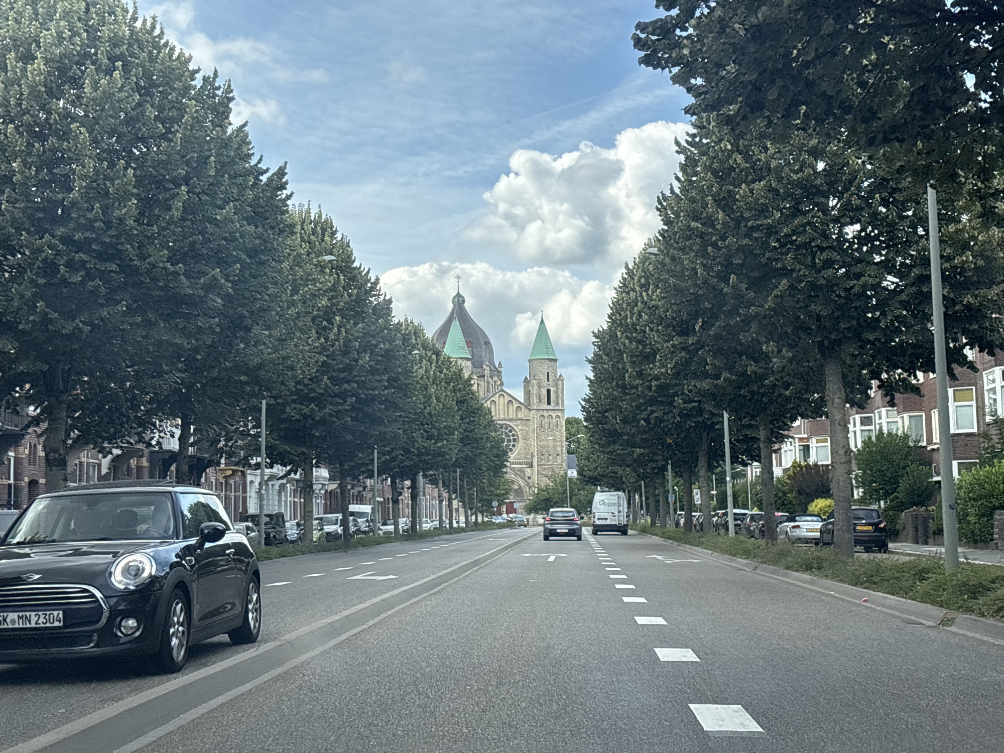 The image shows a street in Maastricht, Netherlands, with a prominent church in the background. The street is lined with mature trees on both sides, creating a canopy over the road. A black Mini Cooper is in the foreground on the left, its license plate reading "GK MN 2304". Several other vehicles are visible on the road and parked along the side. The church, which appears to be a cathedral, features a large dome and spires. The sky is partly cloudy with some cumulus clouds. The architecture of the buildings along the street suggests a European city. The overall scene is one of a peaceful urban environment on a day with moderate weather.
