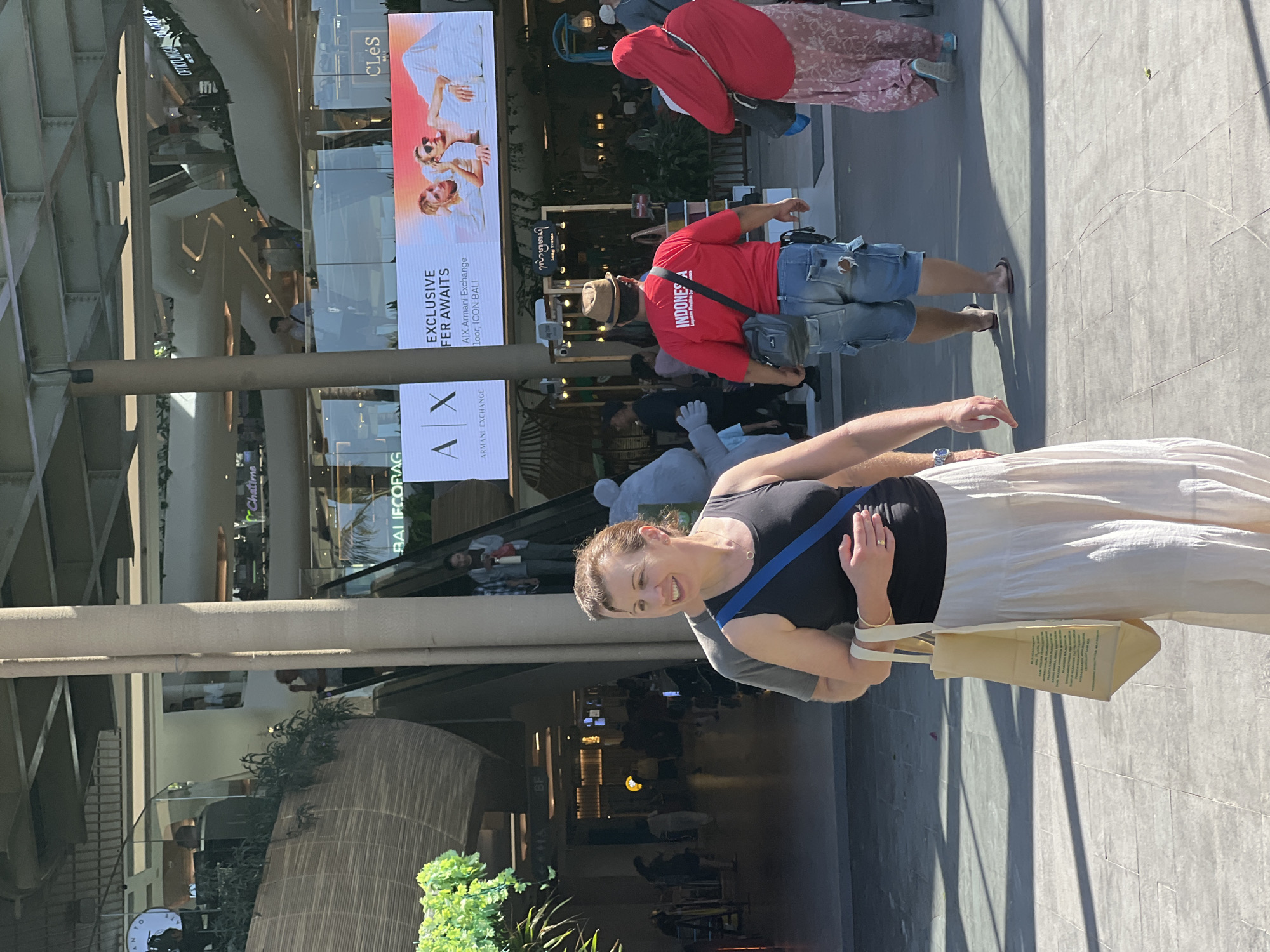 The photograph captures an outdoor scene in Denpasar, Indonesia, likely at a modern shopping complex. In the foreground, a woman wearing a black tank top and a long, light-colored tiered skirt is walking and looking to her left, smiling. She has a blue strap across her chest and is carrying a light-colored tote bag in her right hand. Behind her, a person in a grey shirt is partially visible, appearing to be walking with her.
Further back, a man wearing a red t-shirt with "INDONESIA" printed on it and denim shorts is walking away from the camera, wearing a fedora and a crossbody bag. To his right, a person in a red headscarf and a patterned pink skirt is walking, also away from the camera. Behind them, a large digital advertisement displays "AX ARMANI EXCHANGE" and an image of two people. The environment includes architectural elements of a building with multiple levels, visible escalators, and some greenery. The ground is paved with large square tiles, and shadows suggest bright daylight conditions. The overall atmosphere is that of a busy public space.