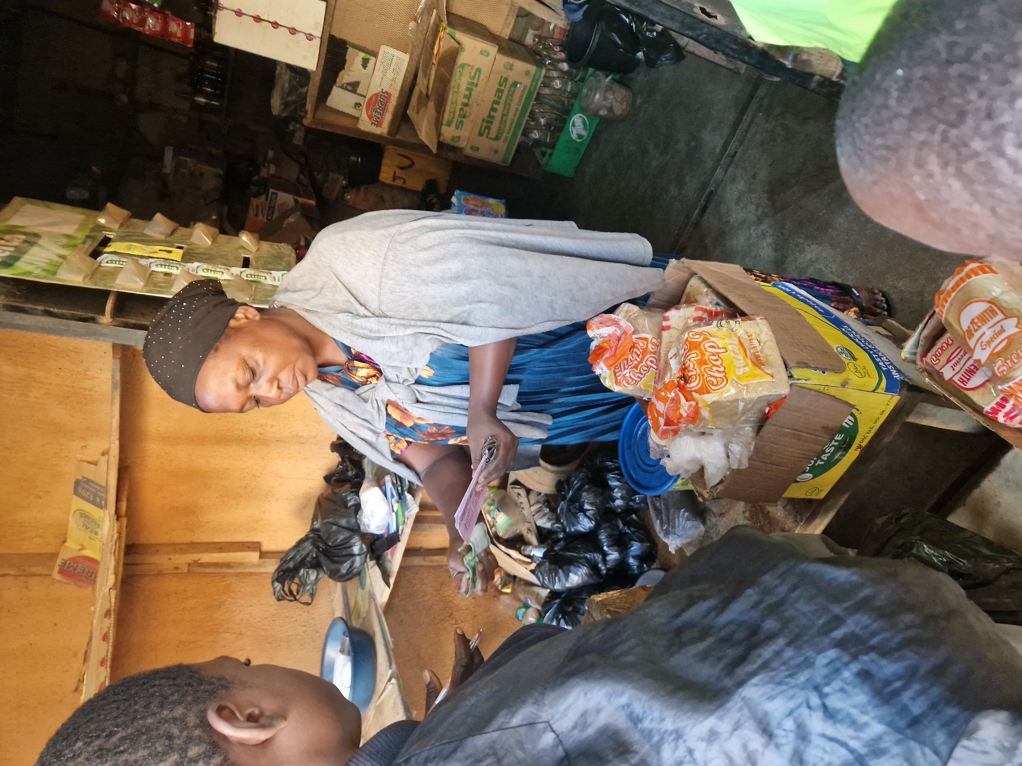 In an indoor market stall, a woman wearing a dark headscarf and a blue dress with an orange and purple floral pattern, draped with a light gray shawl, stands behind a wooden counter. She is holding a fan of banknotes in her right hand and appears to be completing a transaction with another person whose back is to the camera. The stall is stocked with various goods, including bags of bread in a cardboard box in the foreground, plastic bags of what appear to be food items, and shelves in the background displaying bottles and boxes of products. The overall lighting suggests an overcast day or an indoor space with limited natural light.