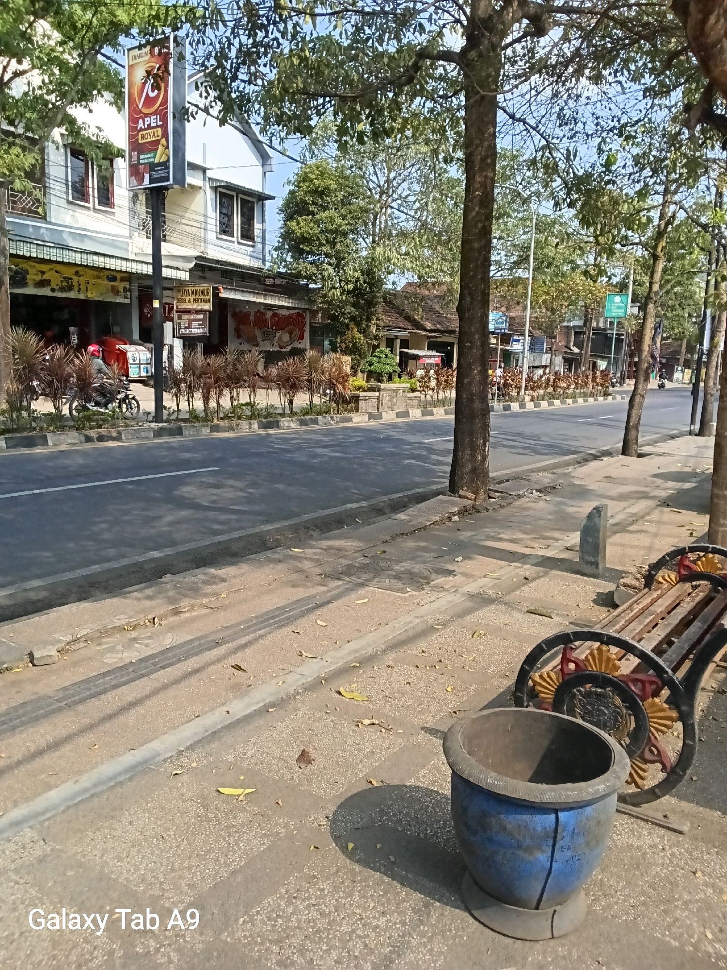 The image captures a street scene in Kelutan, Indonesia, on a sunny day. A paved road runs horizontally across the frame, with a sidewalk and a row of trees lining its right side. The sidewalk features a weathered wooden bench with decorative metalwork and a blue, broken planter. Beyond the road, a two-story building with shops on the ground floor is visible, adorned with signs. One sign promotes "APEL ROYAL." A person wearing a red helmet is riding a motorcycle on the road, and further down the street, a few other individuals are present, along with more buildings and trees. The overall environment suggests a moderately busy urban or suburban area.