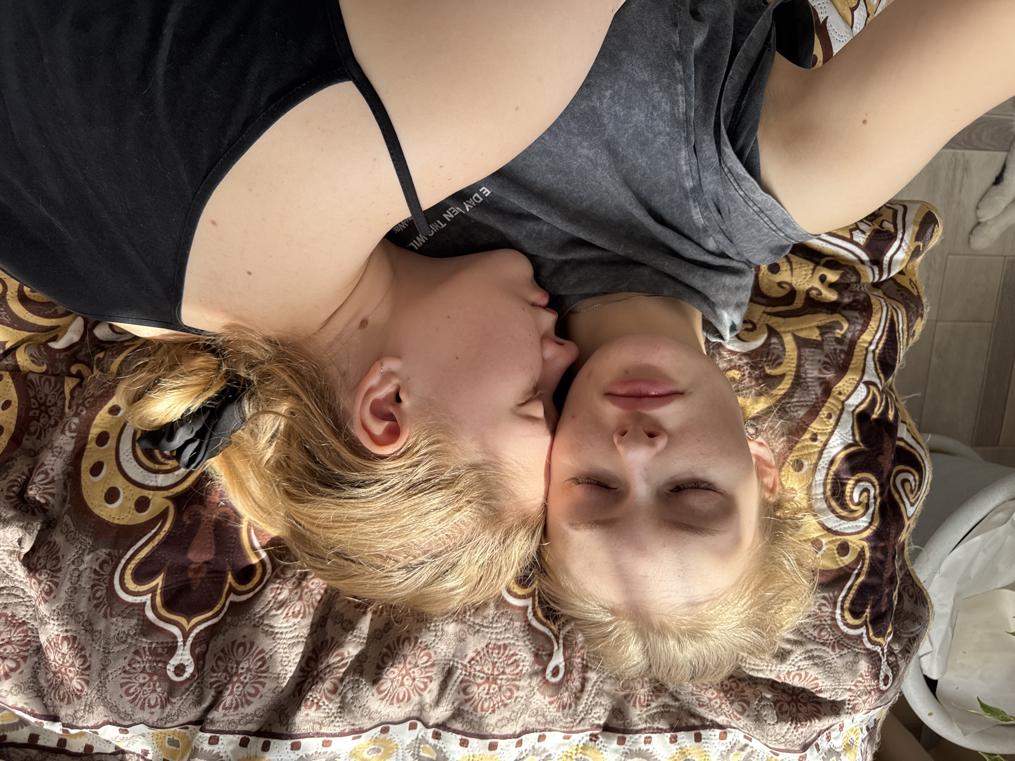 Two fair-skinned women with blonde hair are lying head-to-head on a patterned brown and gold bedspread. The woman on the left is wearing a black tank top and has a dark hair tie in her hair. She is kissing the cheek of the woman on the right, who is wearing a grey t-shirt and has her eyes closed. The location is not evident from the image.