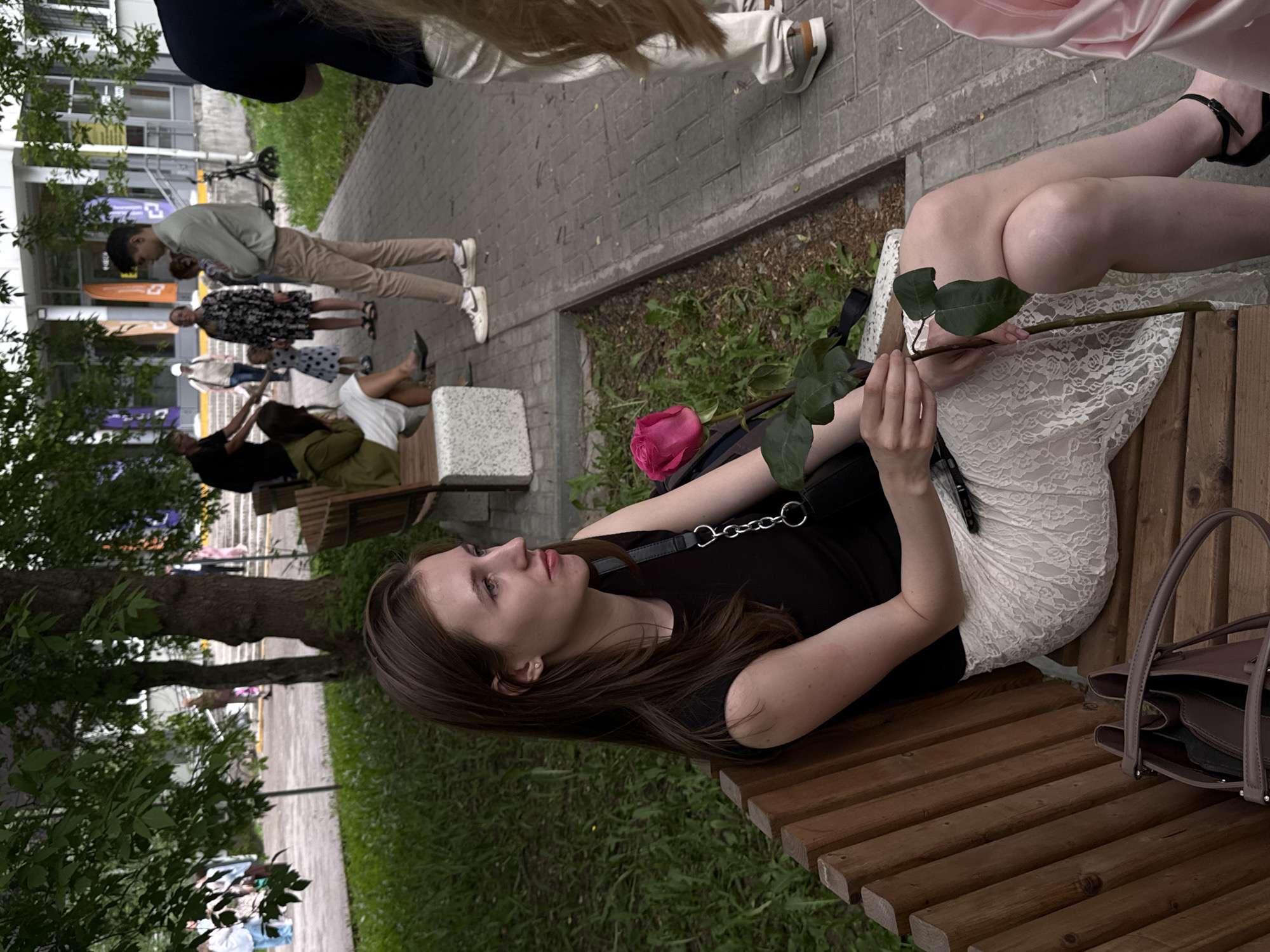 The image shows a young woman sitting on a wooden bench in a park. She has long brown hair and is wearing a black top and a white lace skirt. She is holding a rose. Next to her on the bench is a dark-colored handbag. A pink rose lies on the bench as well. Other people are visible in the background, walking on a paved path in what appears to be a public area. The location is Perm, Russia, based on the architecture and environment typical of the city. There is greenery surrounding the bench, with trees on one side and grass underneath. A sidewalk with people walking on it can be seen in the background, as well as buildings typical of an urban environment.