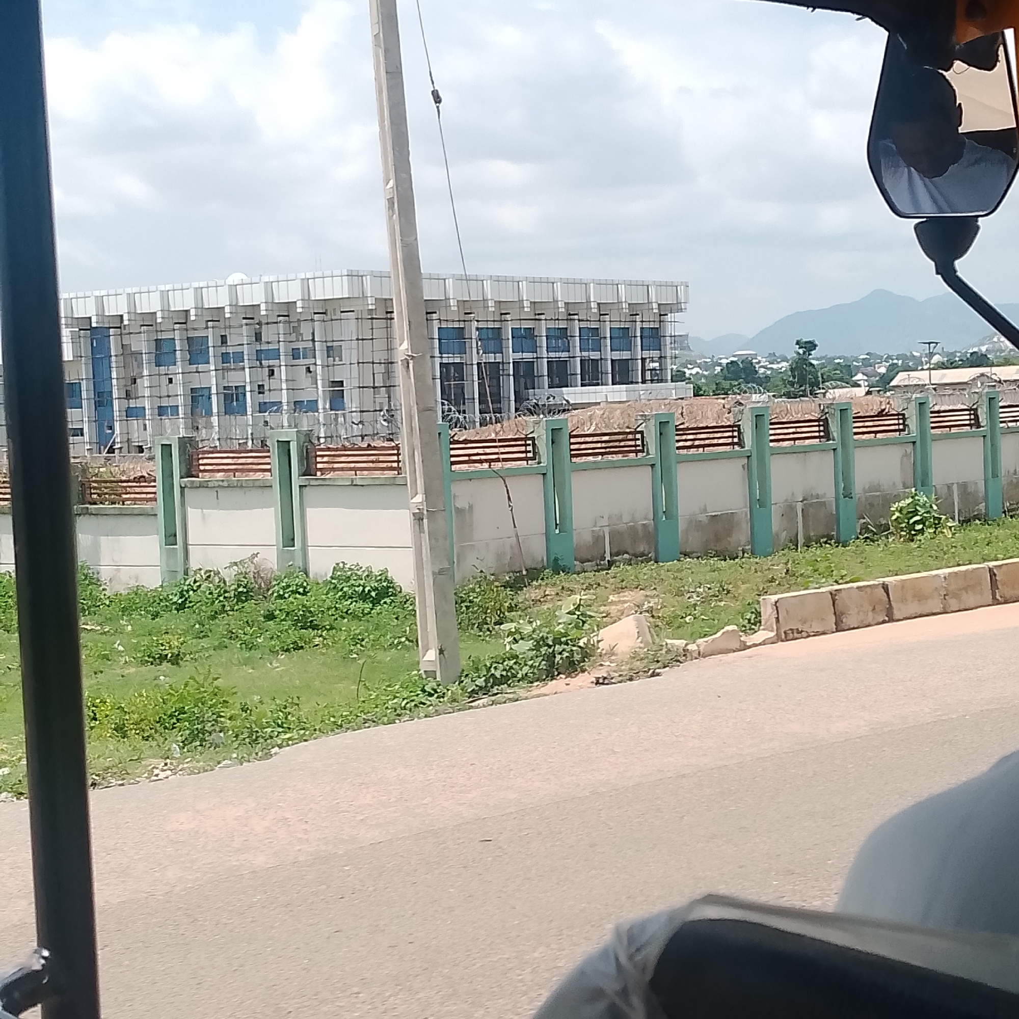 The image shows an exterior view of a modern building under construction, likely in Jalingo, Nigeria. The building features a repetitive facade with numerous windows, suggesting a large office or residential complex. The structure appears to be in the early stages of completion, with visible scaffolding and construction materials. In front of the building, there is a low wall topped with a railing and barbed wire, indicating security measures. Greenery, including grass and some bushes, grows in front of and around the wall, suggesting a somewhat overgrown or naturalistic landscaping. A paved road is visible in the foreground, with a portion of a vehicle, possibly a rickshaw or tuk-tuk, partially obscuring the lower left corner. In the background, hills or mountains are visible, characteristic of a landscape with elevation changes. The sky is partly cloudy, suggesting daytime with fair weather. The overall scene depicts an active construction site in a semi-urban or suburban environment.
