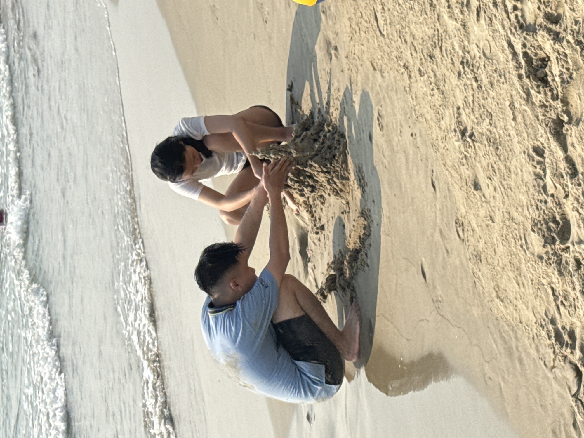 Two people are on a sandy beach constructing something with wet sand. The man is wearing a light blue t-shirt and black shorts. The woman is wearing a white t-shirt and black shorts. The scene is on a beach adjacent to the ocean. Based on the landscape and activities, it is likely in a coastal area such as Đà Nẵng, Vietnam.