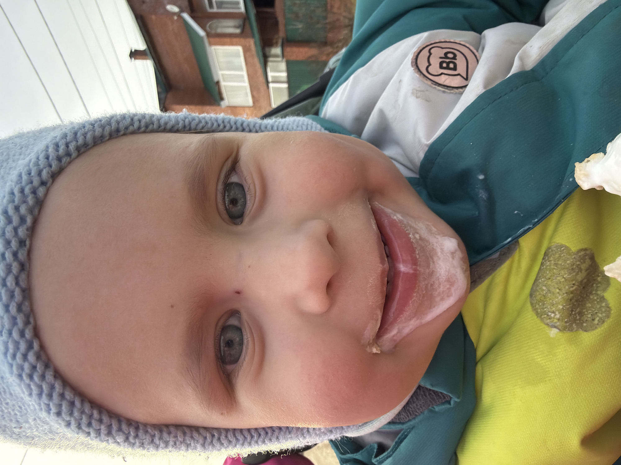 A close-up, slightly rotated, high-angle shot depicts the face of a child, likely in the city of Chaikovsky, Russia. The child is wearing a light blue knit hat. Their eyes are light gray-blue. The child's mouth is slightly open, with their tongue protruding and coated with a white, possibly creamy substance. Traces of the same white substance are visible around their mouth and on the front of their clothing. The clothing appears to be a multi-layered jacket in shades of white, teal, and yellow. A logo with the letters "Bb" is visible on the jacket. A building is partially visible in the background. The environment appears to be outdoors, possibly during the day. A stain is visible on the yellow jacket.
