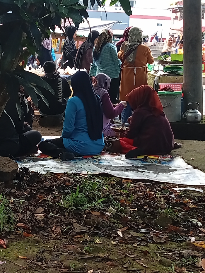 This image captures a vibrant outdoor scene in Singaparna, Indonesia, depicting a communal gathering or market activity. The foreground consists of dry leaves and sparse green vegetation, partially shadowed by a large, leafy plant on the left.
In the mid-ground, several individuals are seated on a colorful mat or tarp spread on the ground. Three women are prominently featured, seen from behind or the side. One woman wears a bright blue top and a long black hijab, seated with her legs crossed. Beside her, another woman in a light purple outfit and a lighter-toned hijab is also seated, engaged with items in front of her. To the right, a woman in a maroon garment and a reddish-brown hijab sits facing away from the camera. They appear to be socializing or consuming food and drink. Further left, a man in a black long-sleeved shirt, with the word "SOUVENIR" faintly visible on the back, is seated in a similar fashion.
The background reveals a bustling market environment with numerous people, primarily women in hijabs, standing around various stalls. Goods, likely food items wrapped in banana leaves or plastic, are displayed on tables and in baskets. A white bucket and a metal teapot are visible near a pillar on the right side of the frame. Buildings with simple architecture can be seen in the far background.
The time of day appears to be daytime, possibly morning or early afternoon, characterized by soft, diffused natural light that suggests an overcast sky or significant shading. The overall atmosphere is relaxed and social, typical of a local community gathering or market.