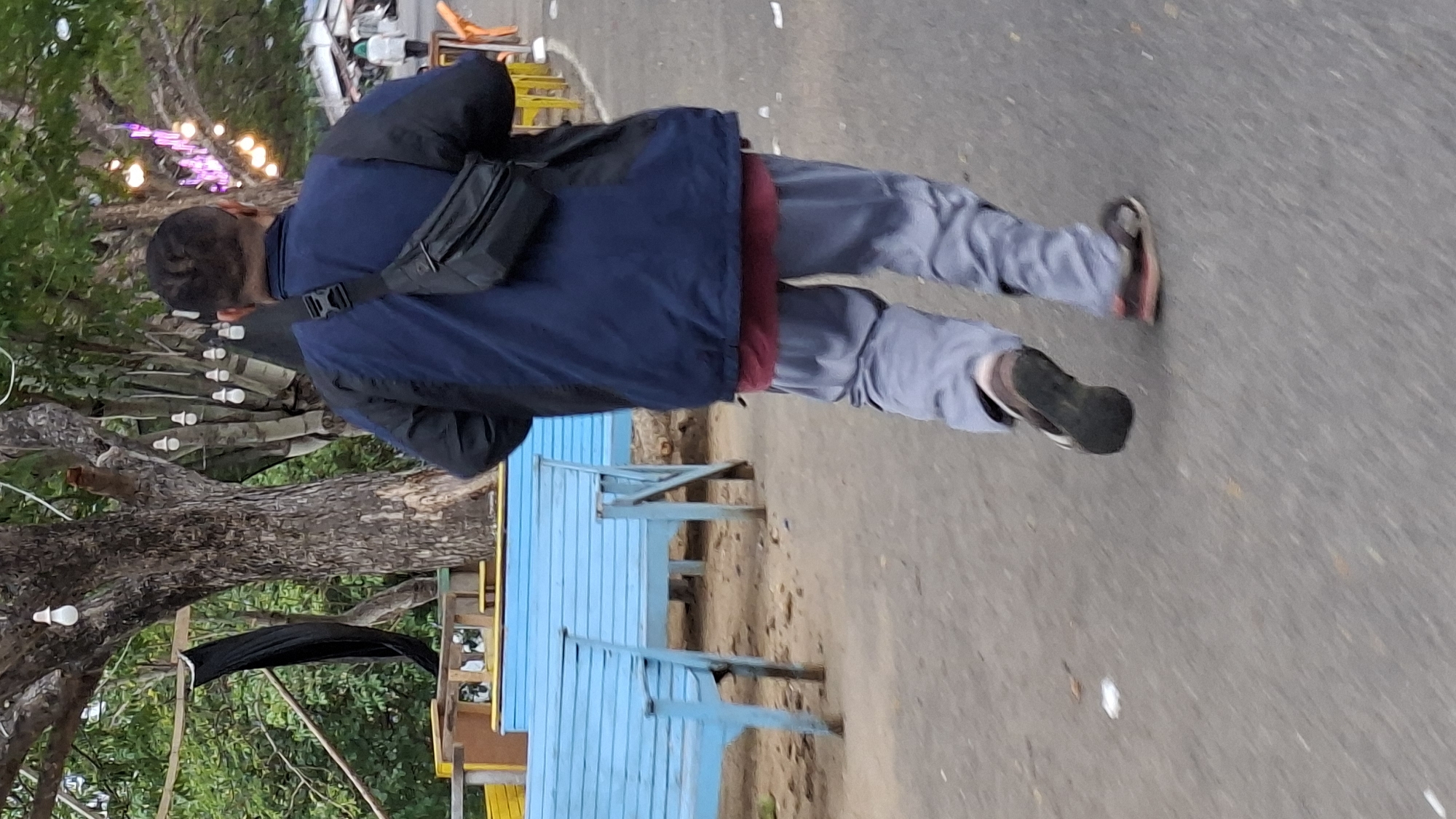 An individual, seen from the rear, walks on a paved surface. The person is wearing a dark blue jacket over a maroon shirt, light grey pants, and sandals. A black fanny pack is worn across the front of the jacket. In the background, several light blue benches are visible. Trees with exposed bark and foliage line the area, with decorative string lights, some illuminated with purple and white bulbs, adorning branches. The scene appears to be an outdoor public space, potentially a park or recreational area within Lhokseumawe, Indonesia.