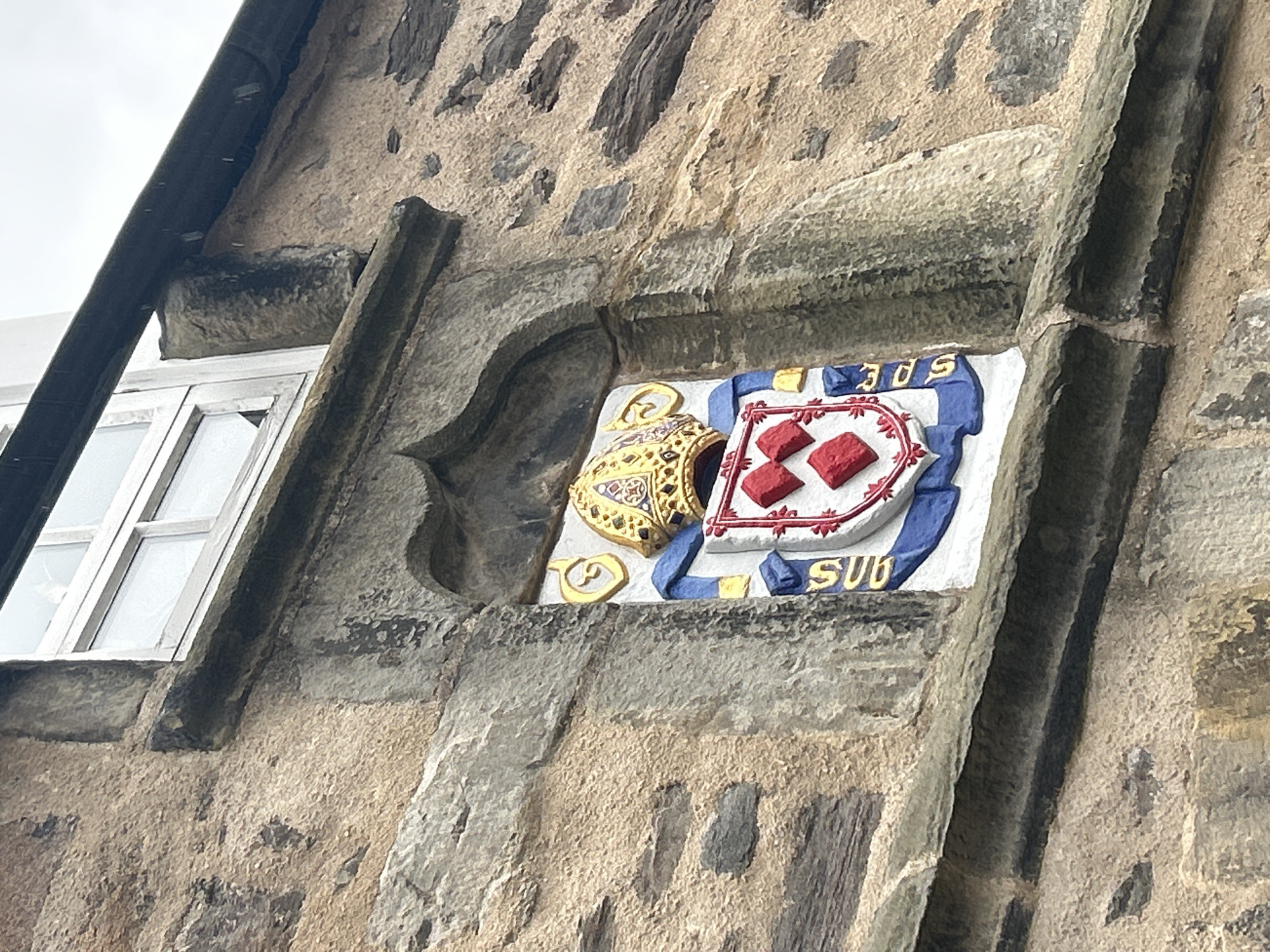 The image shows a heraldic panel set into a stone building, possibly in Aberdeen City, United Kingdom. The panel features a coat of arms with a decorated helmet crest to the left and a shield with a white field bearing three red lozenges within a red border. A blue ribbon-like device surrounds the shield with the letters "SPE" above and "SUB" below. The surrounding building is made of rough, weathered stone, and a window is visible to the left of the panel. The architectural style and materials suggest a historical building. The weather appears overcast.