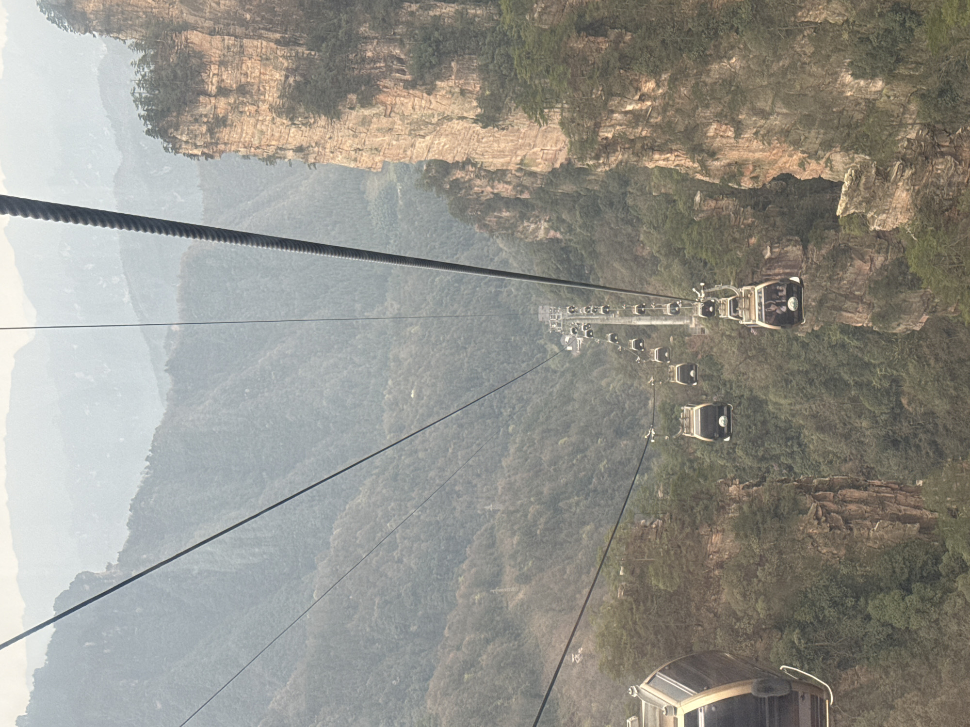 A cable car system descends into a mountainous valley in Wulingyuan, China, viewed from within a moving gondola. Thick, dark gray steel cables extend diagonally across the frame. Approximately twenty white gondola cabins with dark windows and gold-colored trim are visible, suspended from the cables and receding into the distance along the descent line, supported by multiple pylons. Passengers are discernible within at least two of the visible gondolas. To the right, a prominent, vertically striated sandstone cliff face, displaying hues of light brown and orange, rises steeply. The rugged terrain is densely covered in green foliage, including numerous trees and shrubs, particularly on the slopes and cliff tops. Distant mountain ranges are obscured by a pervasive atmospheric haze. The scene depicts a functional transportation system within a natural, rugged landscape.