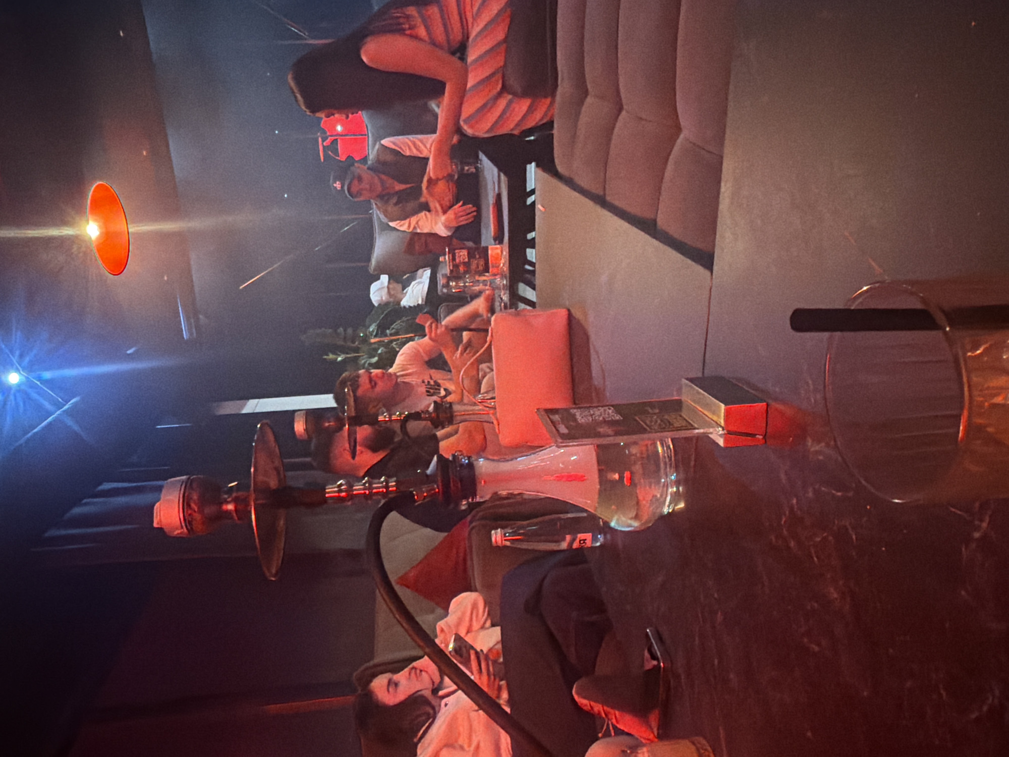 The image depicts an indoor scene in what appears to be a lounge or hookah bar. A group of people are seated on plush seating, some appearing to be interacting while others are engaged with personal devices. A hookah is prominently displayed on a table. Beverages are visible on the table. The location is the city of Varna, Bulgaria.