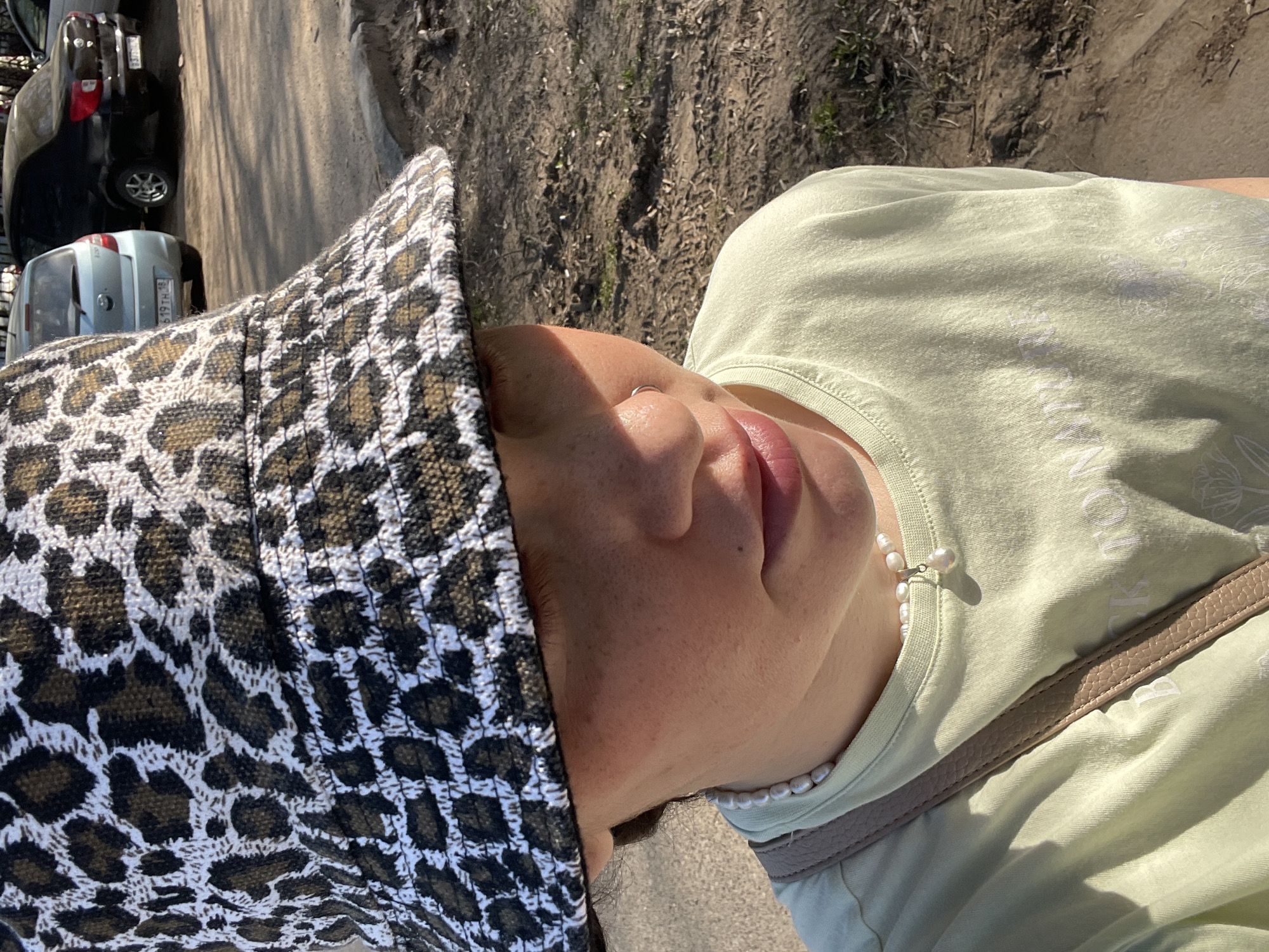A woman is outdoors on a sunny day near what appears to be a street in Izhevsk, Russia. The woman is wearing a light green t-shirt, a string of pearls, and a patterned bucket hat. There are vehicles in the background. The woman is facing upward at an angle, with her face in close view.