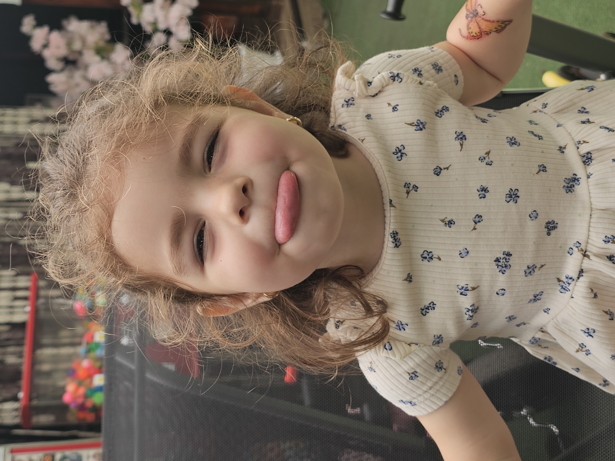 In this close-up shot, a young girl with curly, light brown hair sticks out her tongue, making a playful face. She is wearing a light-colored, short-sleeved dress with a delicate blue floral pattern. A small, gold hoop earring is visible in her left ear, and a colorful butterfly temporary tattoo adorns her right arm. She appears to be seated in an outdoor setting, possibly a park or a backyard, with a blurred background of greenery and some pink flowers. The overall impression is one of lightheartedness and childhood joy. The image does not contain any information about its location in Sofia, Bulgaria.