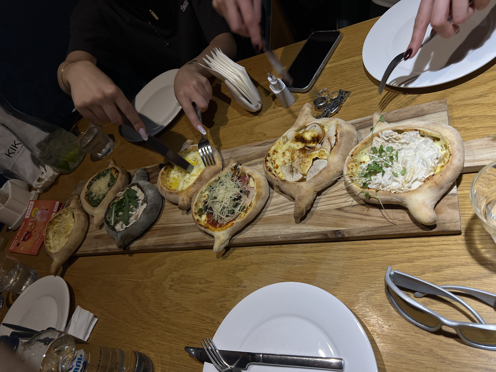 A high-angle, medium shot shows a wooden table laden with several dishes of khachapuri, a Georgian cheese-filled bread, and other food items. A cluster of people are dining in what appears to be a restaurant in Tbilisi, Georgia, evidenced by the presence of a bottle of Borjomi mineral water, a popular Georgian brand.
The khachapuri are served on a wooden board, with several varieties visible. One is topped with spinach and cheese, another with shredded cheese, and one appears to be a classic Adjaruli khachapuri with an egg and butter. There are also plates with silverware, sunglasses, a phone, and napkins. Two women are actively eating, using forks and knives, their hands and fingernails visible. One woman has long, manicured nails and wears a gold bracelet. The other woman's hands are also visible as she reaches for food.
The lighting suggests an indoor setting, possibly a dimly lit restaurant, with the warm glow of the wooden table being prominent. The overall scene depicts a shared meal, likely a social gathering, focused on enjoying traditional Georgian cuisine.