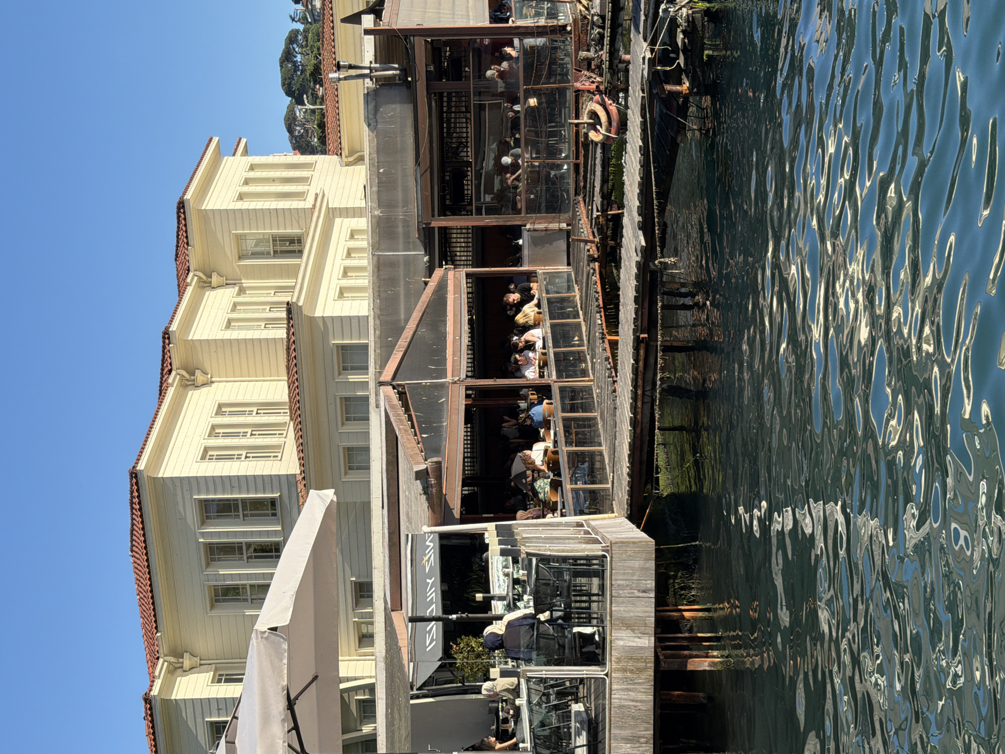 A waterfront scene in Üsküdar, Turkey, features a large, traditional wooden building with a red-tiled roof situated behind a contemporary, open-air restaurant complex built on stilts over the water. The restaurant has a glass-enclosed section and an open dining area with numerous tables and chairs occupied by patrons. In the foreground, the dark, rippling water reflects the sunlight, creating an abstract pattern of light and shadow. The sky is a clear, bright blue. The restaurant appears to be a popular dining spot, with many people seen eating and socializing at tables. A white umbrella is positioned to provide shade for one of the outdoor seating areas. The wooden structure of the restaurant extends out over the water, with a wooden walkway connecting different sections.