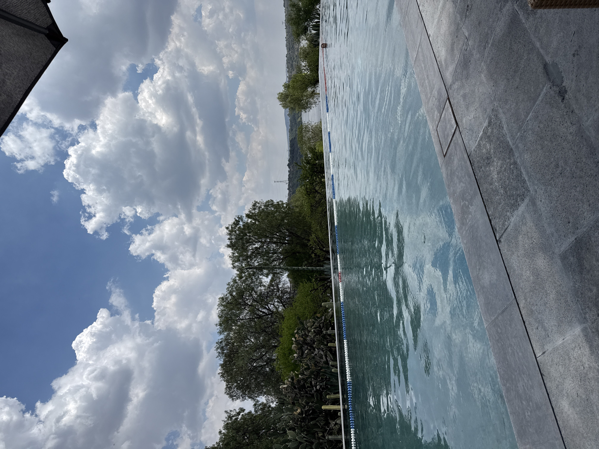 The image shows a view of a swimming pool located in Pueblo Nuevo de Jasso, Mexico, bordered by stone tiles on one side and a line of trees and cacti on the other. A lane divider floats in the water. The sky is partly cloudy with blue patches. In the distance, hills and a transmission tower are visible. The perspective is slightly tilted. A gray structure appears in the upper left corner of the frame.