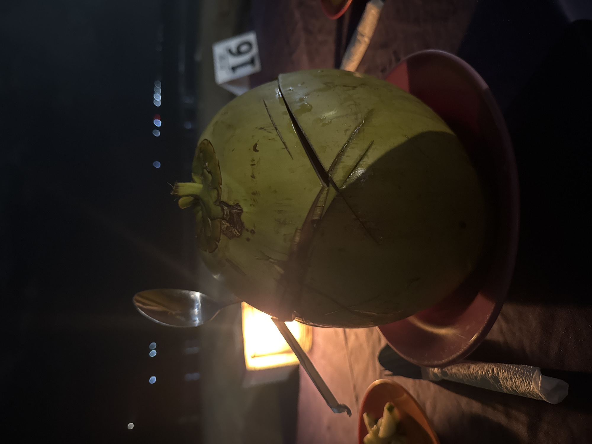 A whole green coconut, scored with decorative lines, sits on a reddish-pink plate on a dark tablecloth. A spoon is inserted into the top of the coconut, and a straw emerges from the side. In the background, a square lantern emits a warm glow, and a table number "16" is visible. The scene appears to be outdoors at night, with distant lights suggesting a beach or coastal area, possibly in Kuta Selatan, Indonesia.