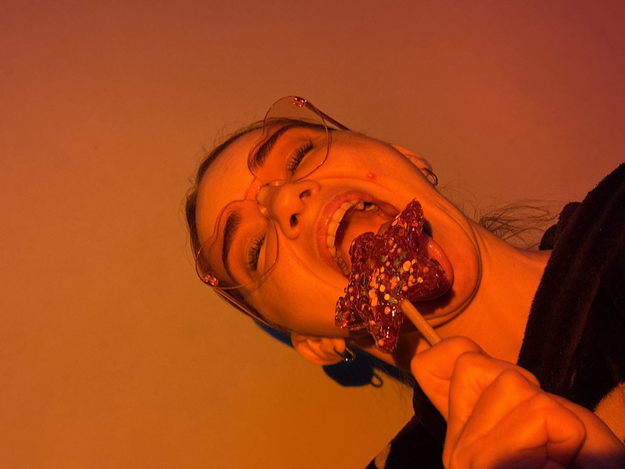 A person with closed eyes and open mouth is holding a red glittery star-shaped lollipop in their mouth. They are wearing heart-shaped glasses and a dark colored top. The background is a gradient of orange and beige. The location is Unknown, Russia.
