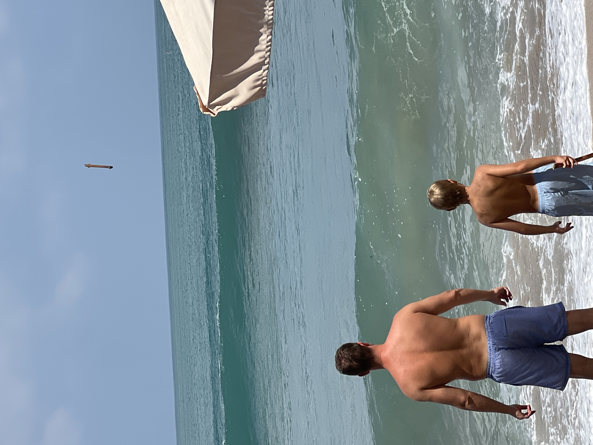 A man and a boy stand on a sandy beach facing the ocean. The man is shirtless and wearing blue striped shorts. The boy is also shirtless and wearing blue shorts. They are looking out at the water where waves are breaking. To the right of the frame, a portion of a tan beach umbrella is visible. In the sky above the ocean, a long, thin object is suspended. The sky is clear and blue. The location is identified as Kuta, Indonesia.