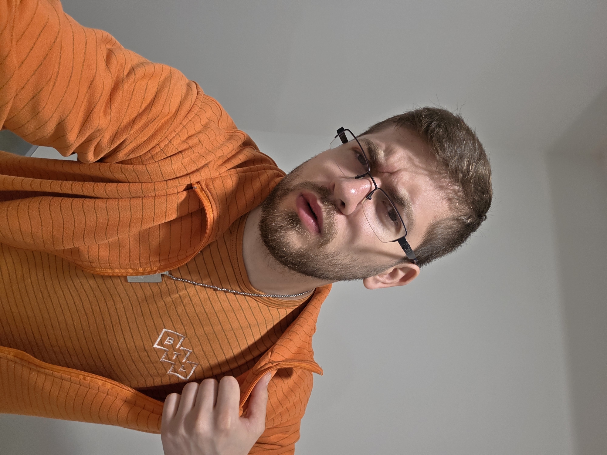 A man wearing an orange zip-up hoodie and an orange shirt with an emblem is looking down at the camera with a furrowed brow. He has short brown hair, a beard, and is wearing glasses. He also appears to be wearing a chain necklace. The background is a neutral gray wall. There is no evidence to suggest that this picture was taken in Plovdiv, Bulgaria.