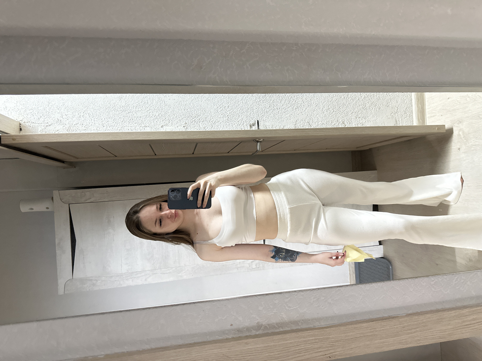 A young woman is captured in a mirror selfie, standing indoors in a room with light-colored decor. She has long, light brown hair and a neutral expression, looking at the black smartphone held in her left hand, which features a triple-camera setup. She is wearing a matching two-piece ribbed outfit in a light color, likely white or off-white, consisting of a crop top and wide-leg pants.
A prominent tattoo depicting a dark animal head with blue details is visible on her left forearm. In her right hand, she holds a folded yellow object, possibly a cleaning cloth or sponge. The mirror reflects a light-toned, textured wall and wooden shelving or cabinetry above and below the woman. A light-colored wooden floor is visible at the bottom of the reflection. In the top left of the reflected image, a cylindrical white object is mounted on the wall, and a grey object is partially visible below her right hand. The setting appears to be a modern, casual indoor space.