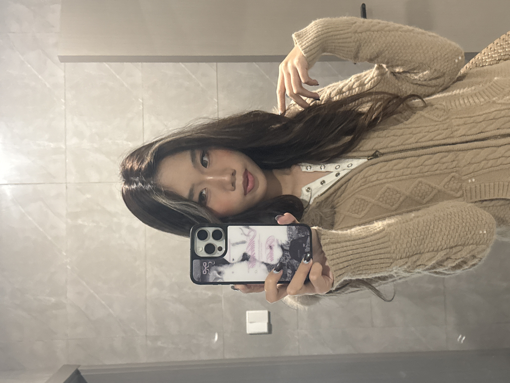 A young female subject, of East Asian descent with long, dark brown hair featuring a blonde streak on the viewer's left, is depicted taking a mirror selfie in Nan'an District, China. She is wearing a light beige cable-knit cardigan over a white snap-button top. Her left hand, with fingernails painted dark with metallic tips, holds a black smartphone with a three-lens camera and a white and pink patterned case, pointed towards her reflection. Her right hand rests on her left shoulder. The background features light grey rectangular wall tiles with a subtle pattern. A white rectangular object is mounted on the wall to the left, and a vertical light grey partition is visible on the right.